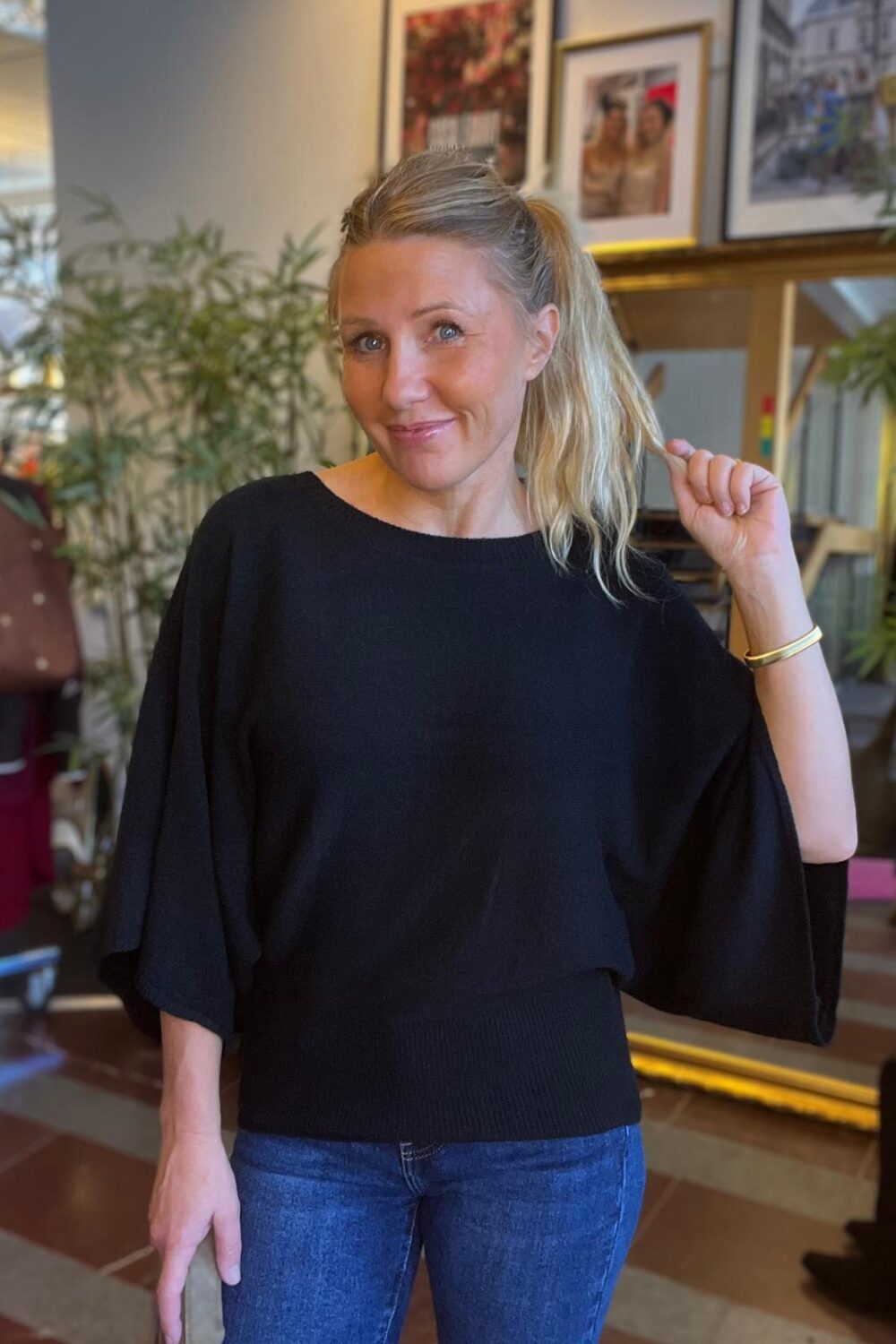 Leende kvinna med blond hästsvans står inomhus och håller i sin hästsvans med ena handen. Hon har svart, löst sittande tröja och blå jeans; i bakgrunden syns krukväxter och inramade fotografier.