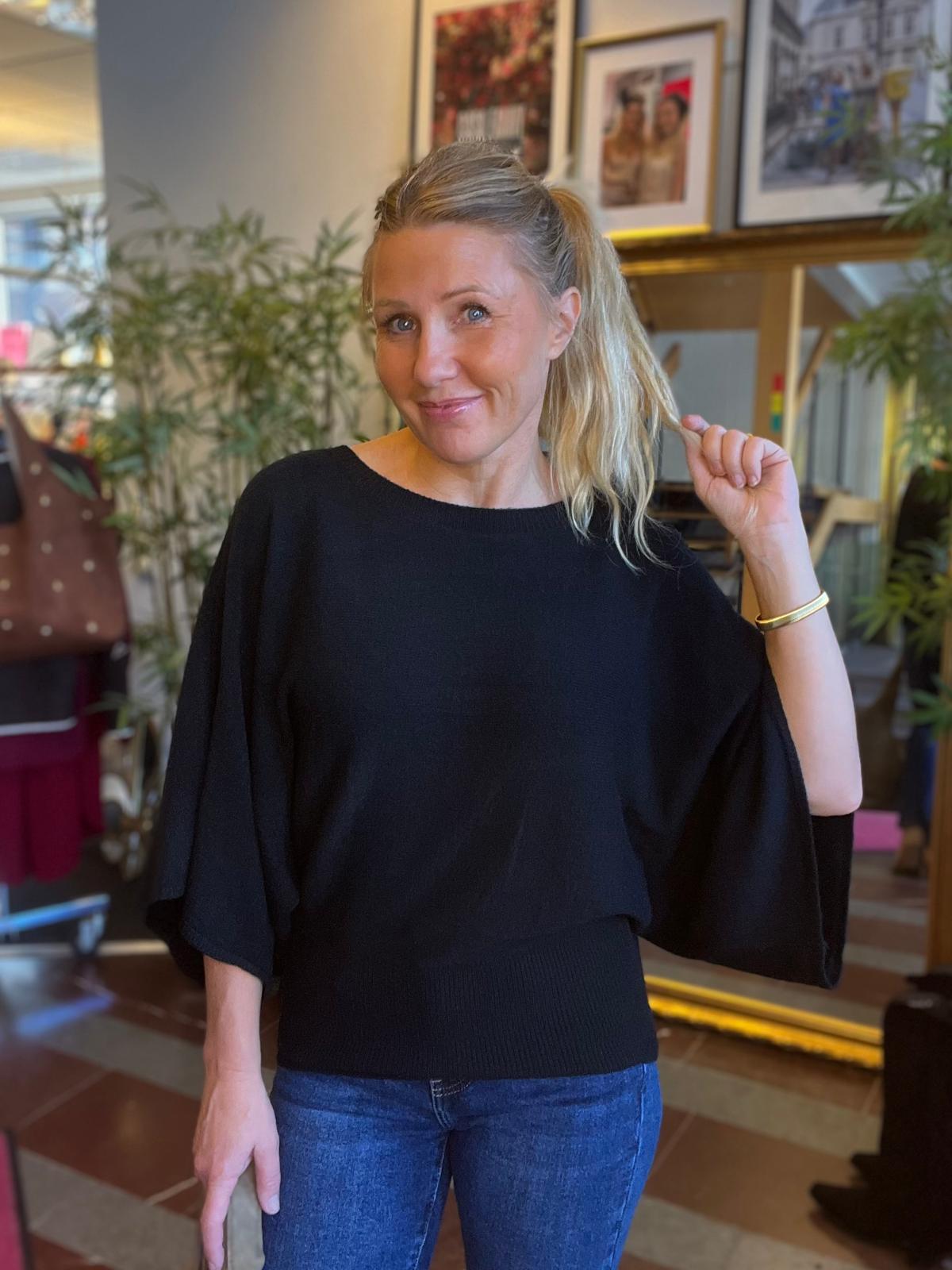 Vaaleahiuksinen nainen poseeraa sisätilassa, hymyilee kameralle ja pitää sivulle vedettyä poninhäntää kädessään. Hänellä on musta leveähihainen paita ja farkut; taustalla näkyy viherkasveja ja kehystettyjä valokuvia.