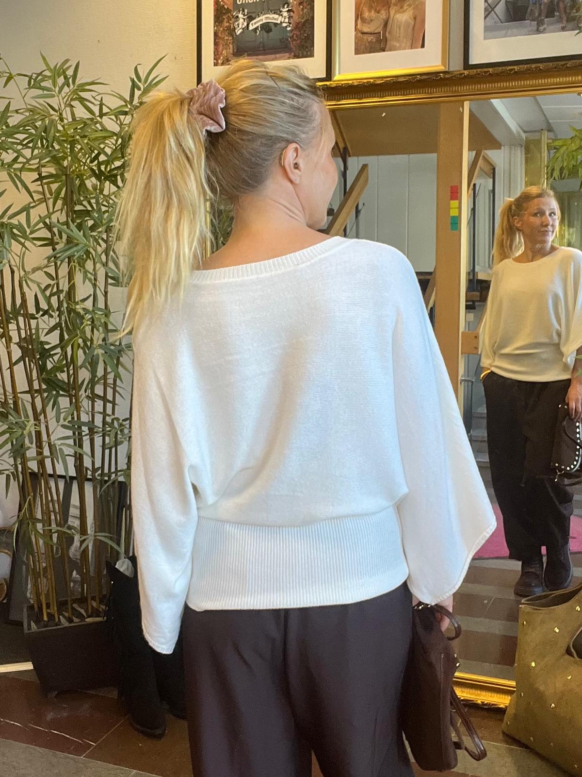 Vaalea, poninhäntäinen nainen seisoo sisätilassa selkä kameraan päin ja katsoo isoon, kultakehyksiseen peiliin. Hänellä on valkoinen neule ja tummat housut, kädessä laukku; taustalla bambukasvi ja kehystettyjä kuvia.