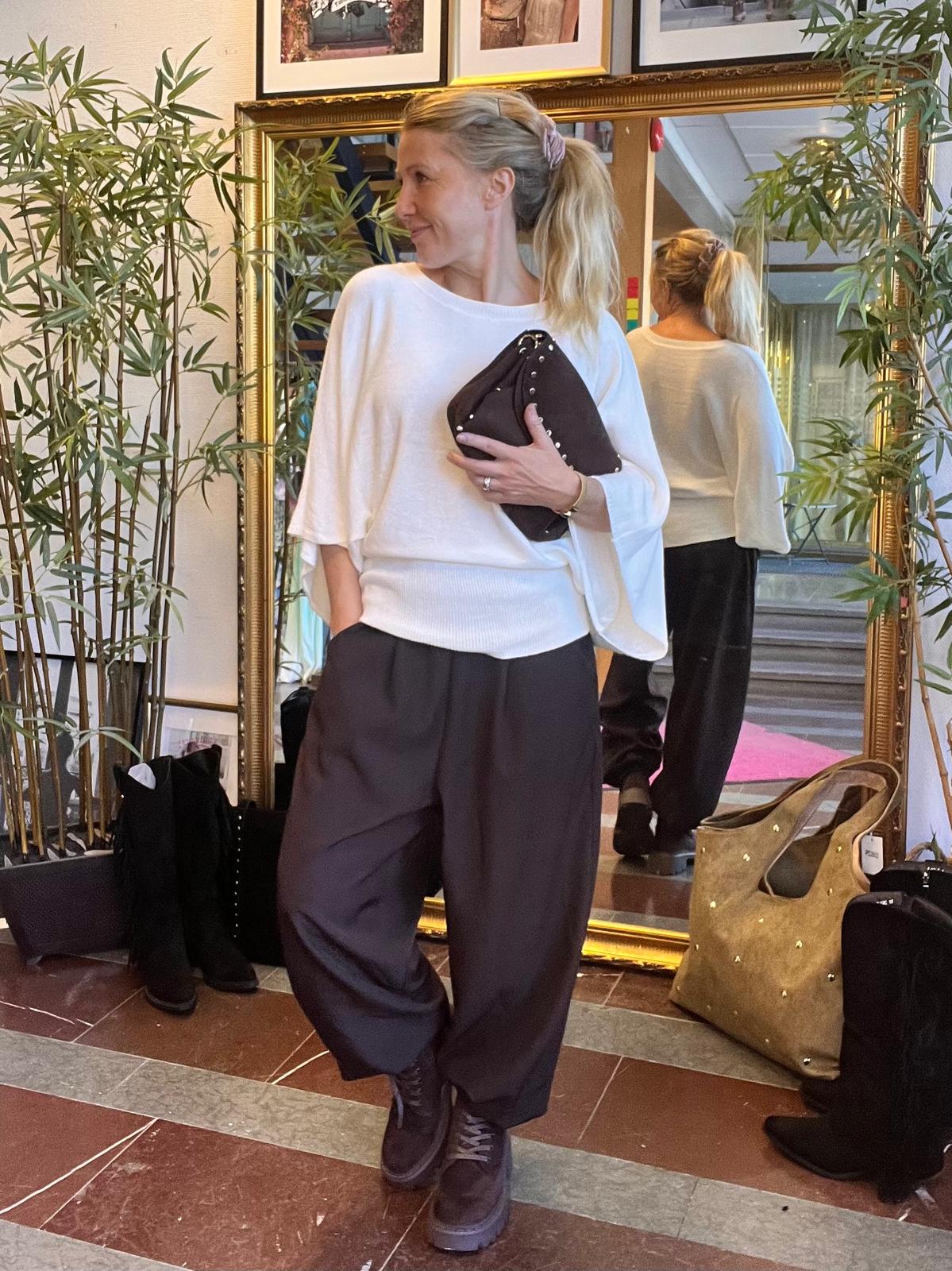 Vaaleahiuksinen nainen poseeraa sisätilassa peilin edessä, katsoo sivulle ja pitää mustaa niiteillä koristeltua clutch-laukkua. Hänellä on valkoinen paita ja tummat väljät housut; peilissä näkyy takaosa, ympärillä bambukasveja ja laukkuja.