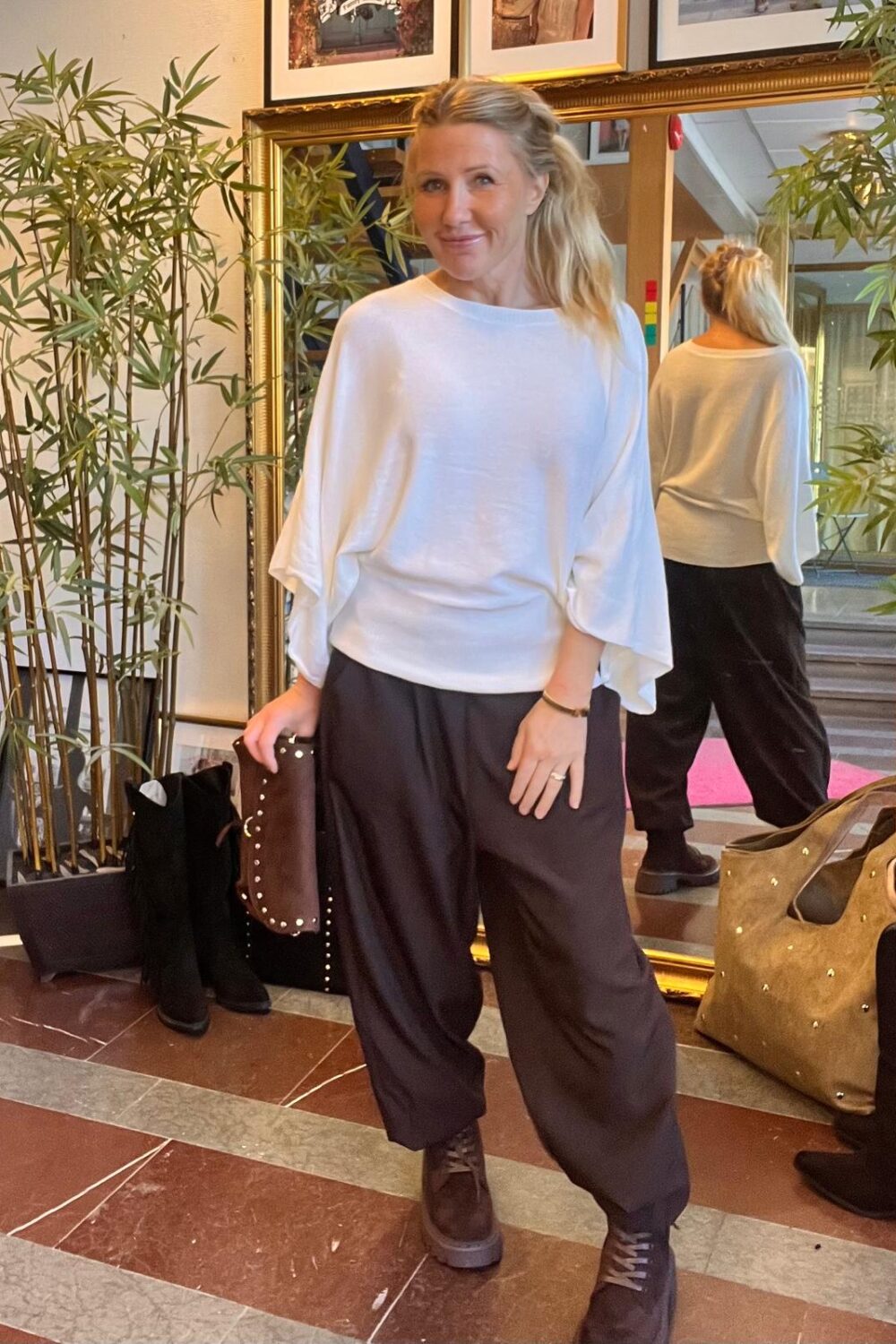 Leende kvinna poserar framför en stor guldspegel och håller en brun nitarad clutch. Hon bär vit, lös topp med vida ärmar, svarta vida byxor och svarta boots, i en hall med bambuväxter och väskor.