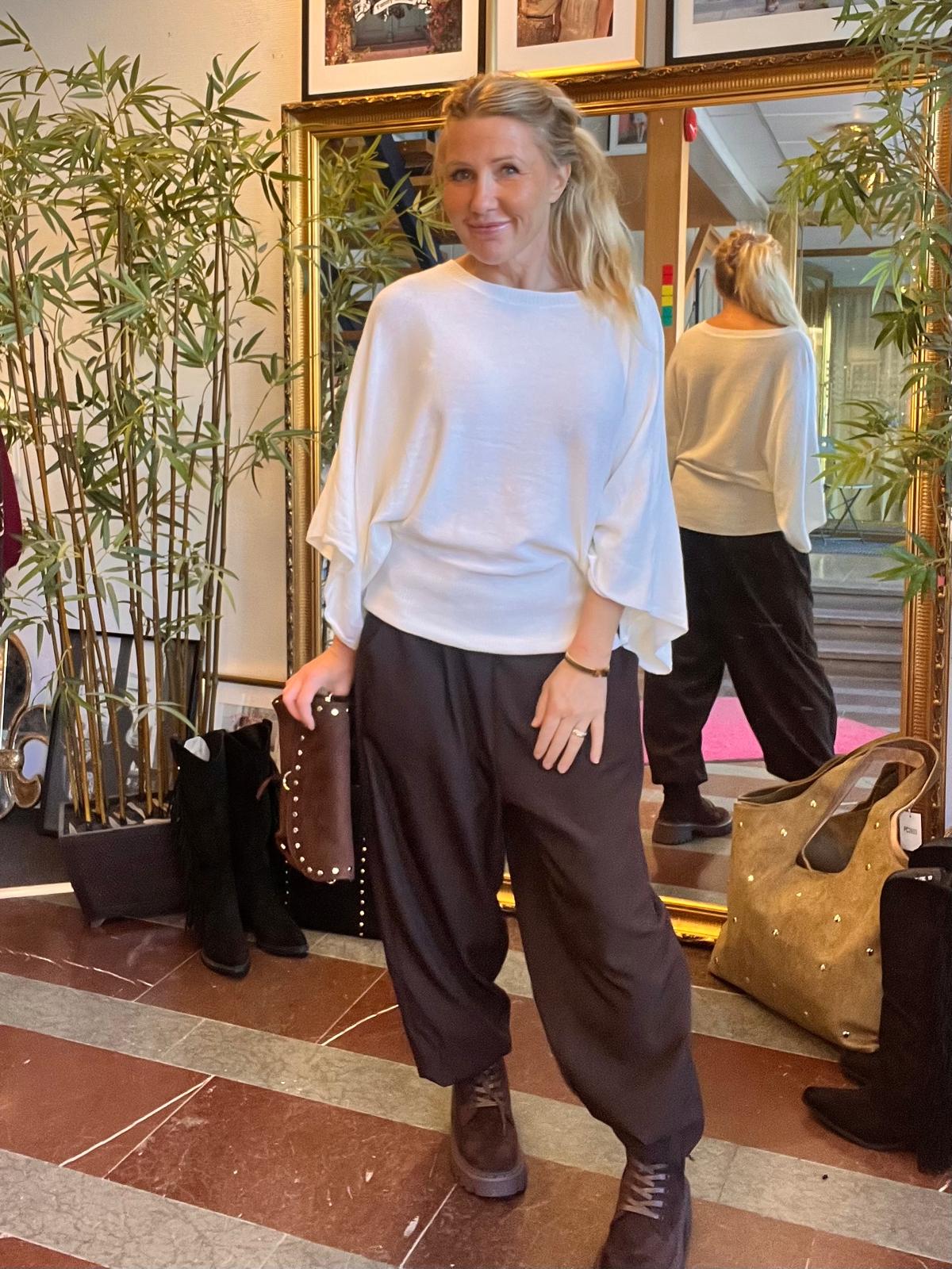Leende kvinna poserar framför en stor guldspegel och håller en brun nitarad clutch. Hon bär vit, lös topp med vida ärmar, svarta vida byxor och svarta boots, i en hall med bambuväxter och väskor.