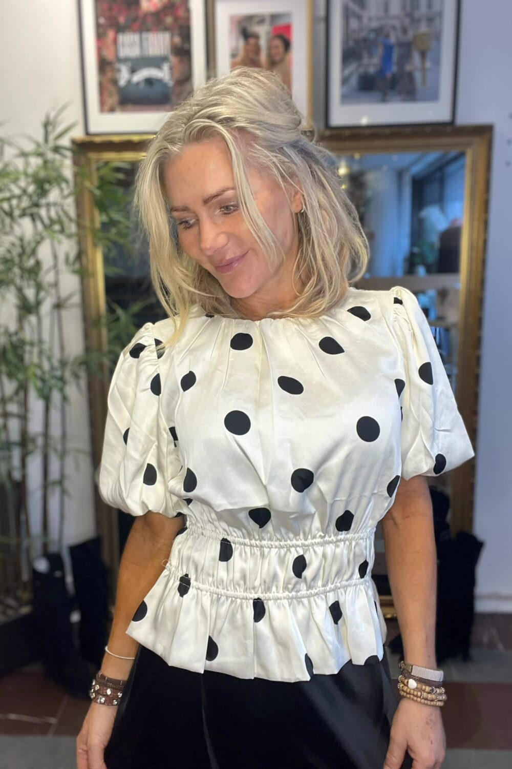 Blond kvinna står inomhus och ler medan hon tittar nedåt, iklädd vit blus med stora svarta prickar och puffärmar samt svart kjol/byxor. I bakgrunden syns spegel, inramade foton och en grön växt.