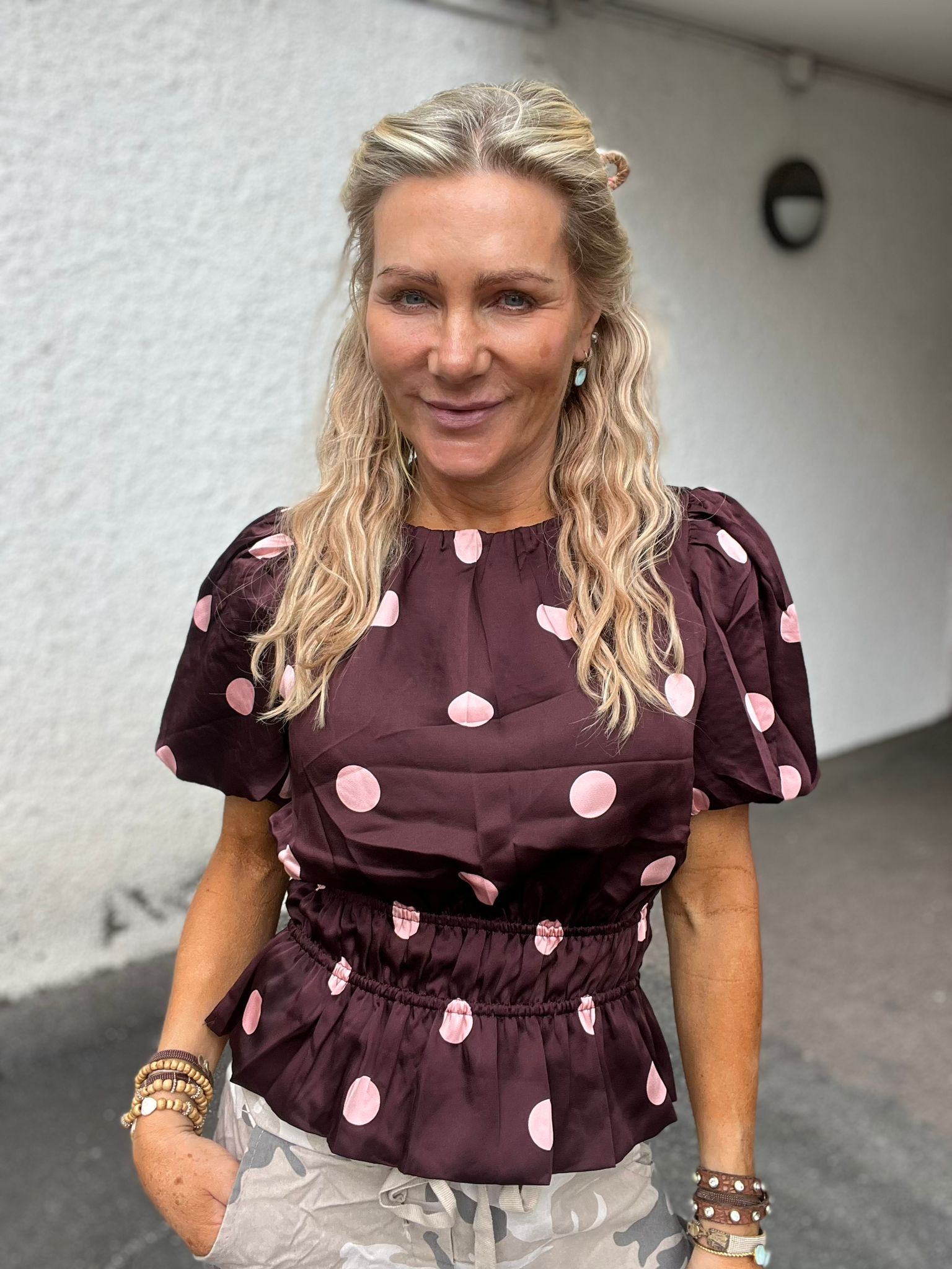 Leende kvinna står utomhus framför en ljus vägg och tittar in i kameran. Hon har långt blont, vågigt hår och bär en vinröd blus med stora rosa prickar samt flera armband.