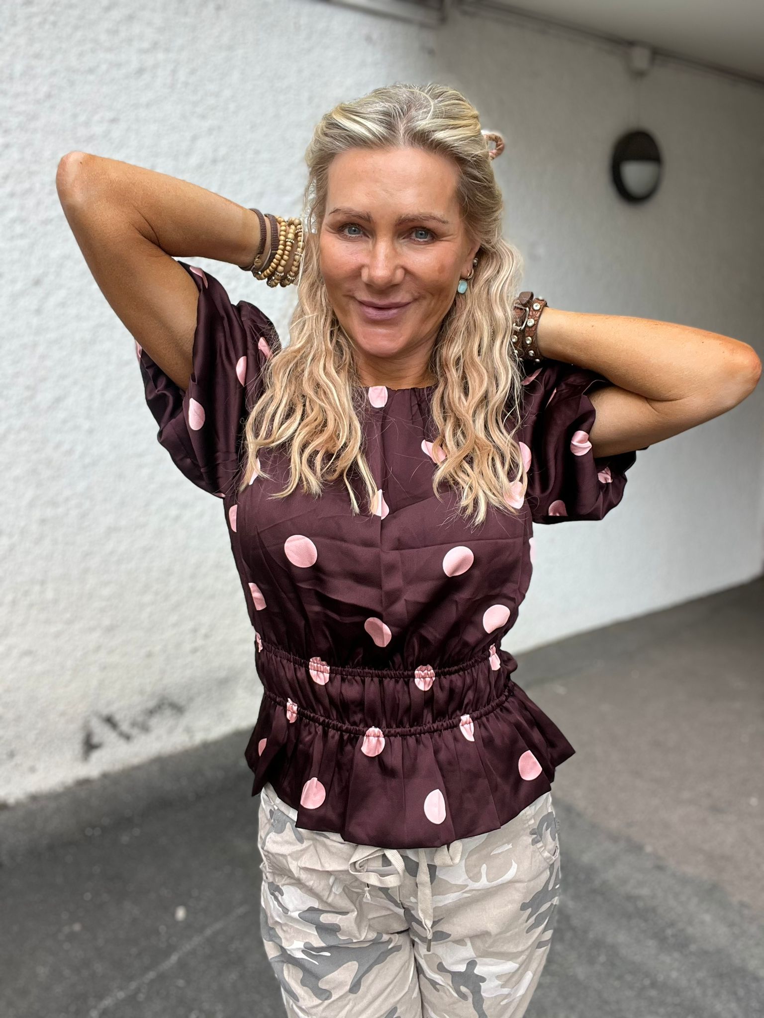 Leende kvinna med långt blont, vågigt hår står med händerna bakom huvudet. Hon bär en mörk blus med rosa prickar, många armband och ljusa kamouflagebyxor, framför en grå vägg utomhus.