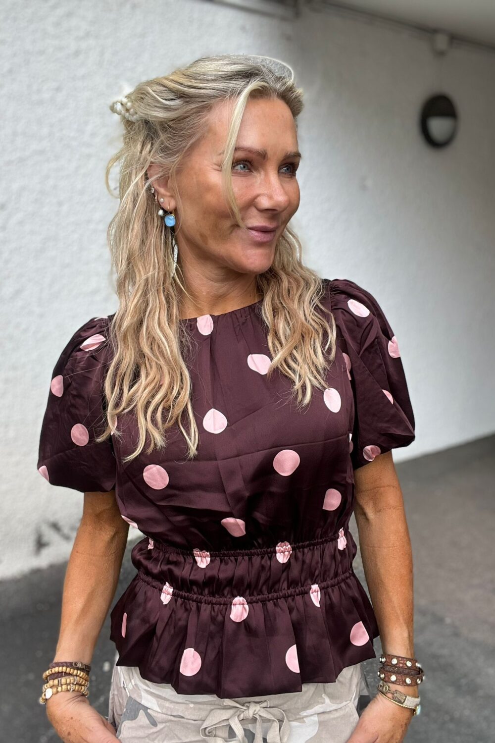Blond kvinna står utomhus och ler medan hon tittar åt sidan. Hon bär vinröd blus med stora rosa prickar, pärlklämma i håret, blå örhänge och många armband, framför en vit vägg med en rund lampa.