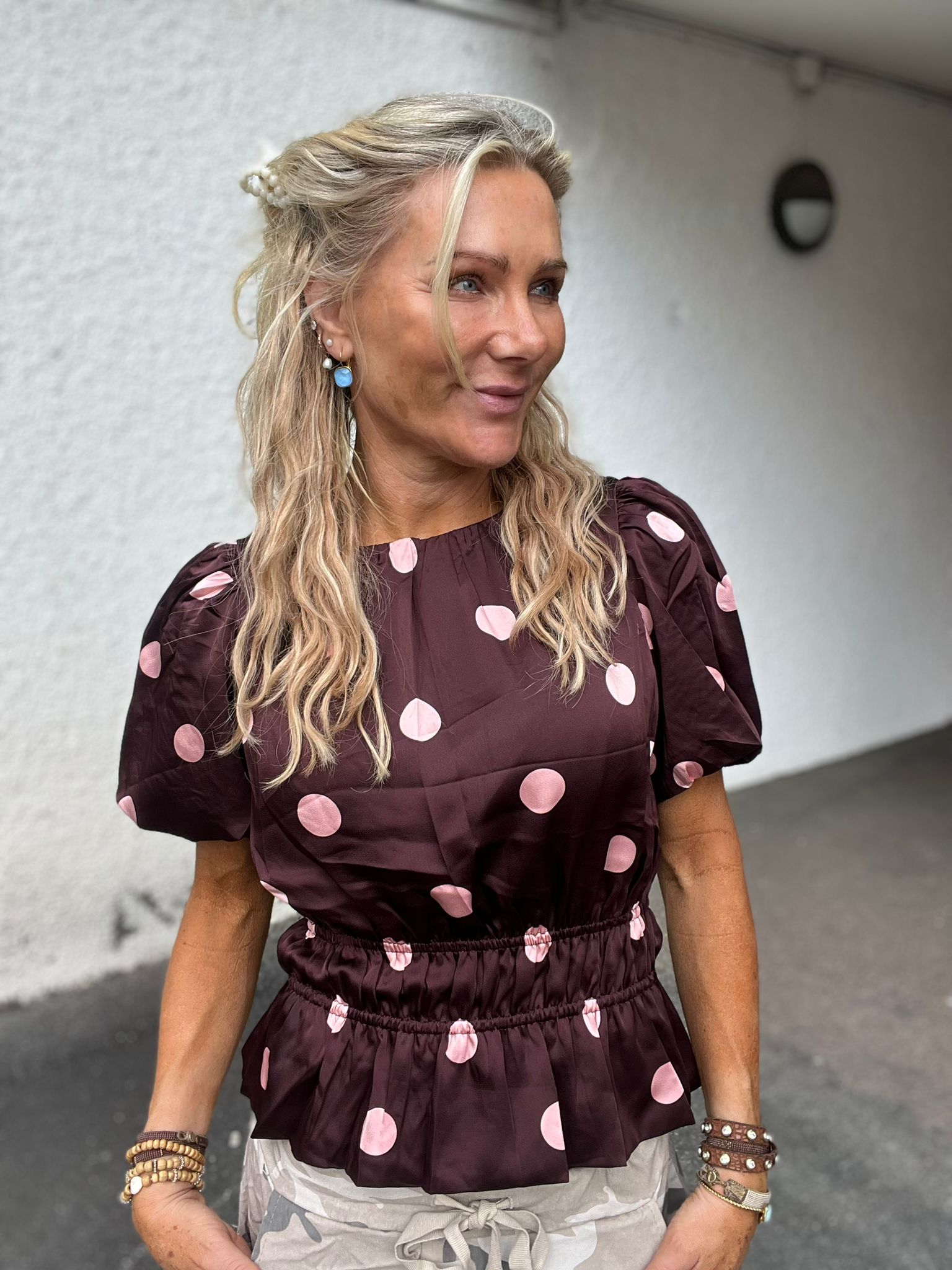 Blond kvinna står utomhus och ler medan hon tittar åt sidan. Hon bär vinröd blus med stora rosa prickar, pärlklämma i håret, blå örhänge och många armband, framför en vit vägg med en rund lampa.