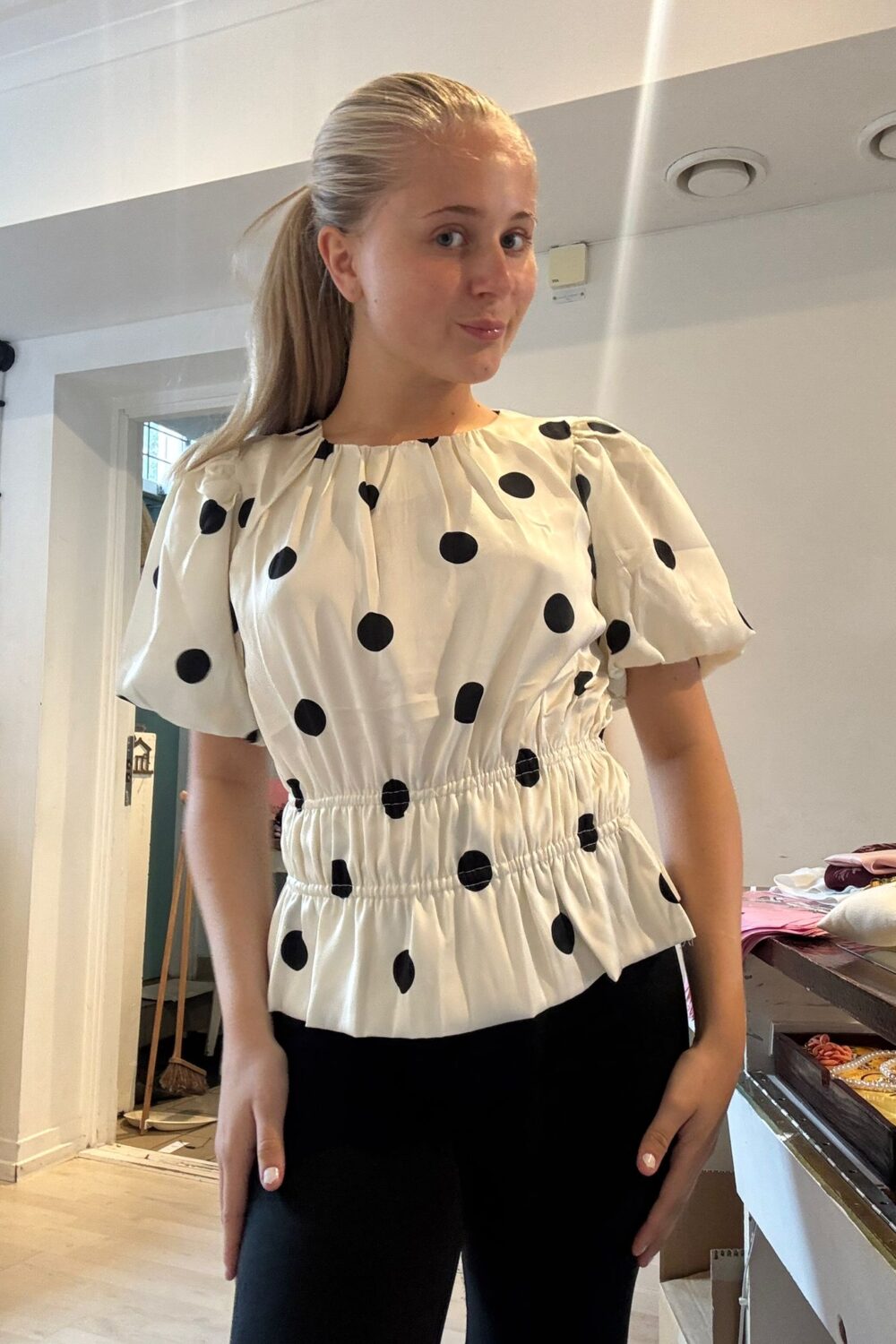 Ung kvinna med blond hästsvans poserar och tittar mot kameran inomhus. Hon bär en vit blus med stora svarta prickar och svarta byxor; i bakgrunden syns ett bord med smycken och andra föremål.