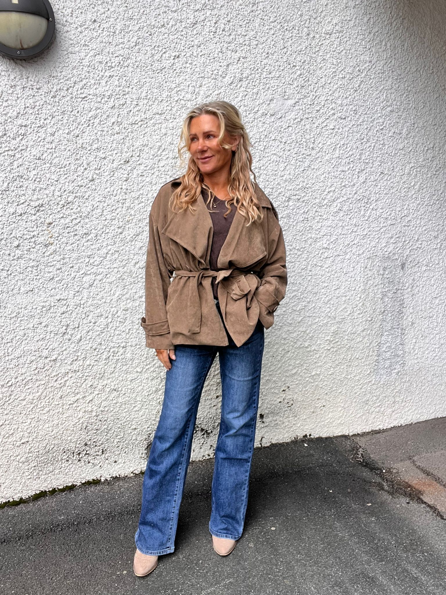 Kvinna med långt blont hår står avslappnat och ler, med ena handen i fickan och blicken åt sidan. Hon bär brun knuten jacka, jeans och ljusa skor, framför en vit grov putsad vägg utomhus.