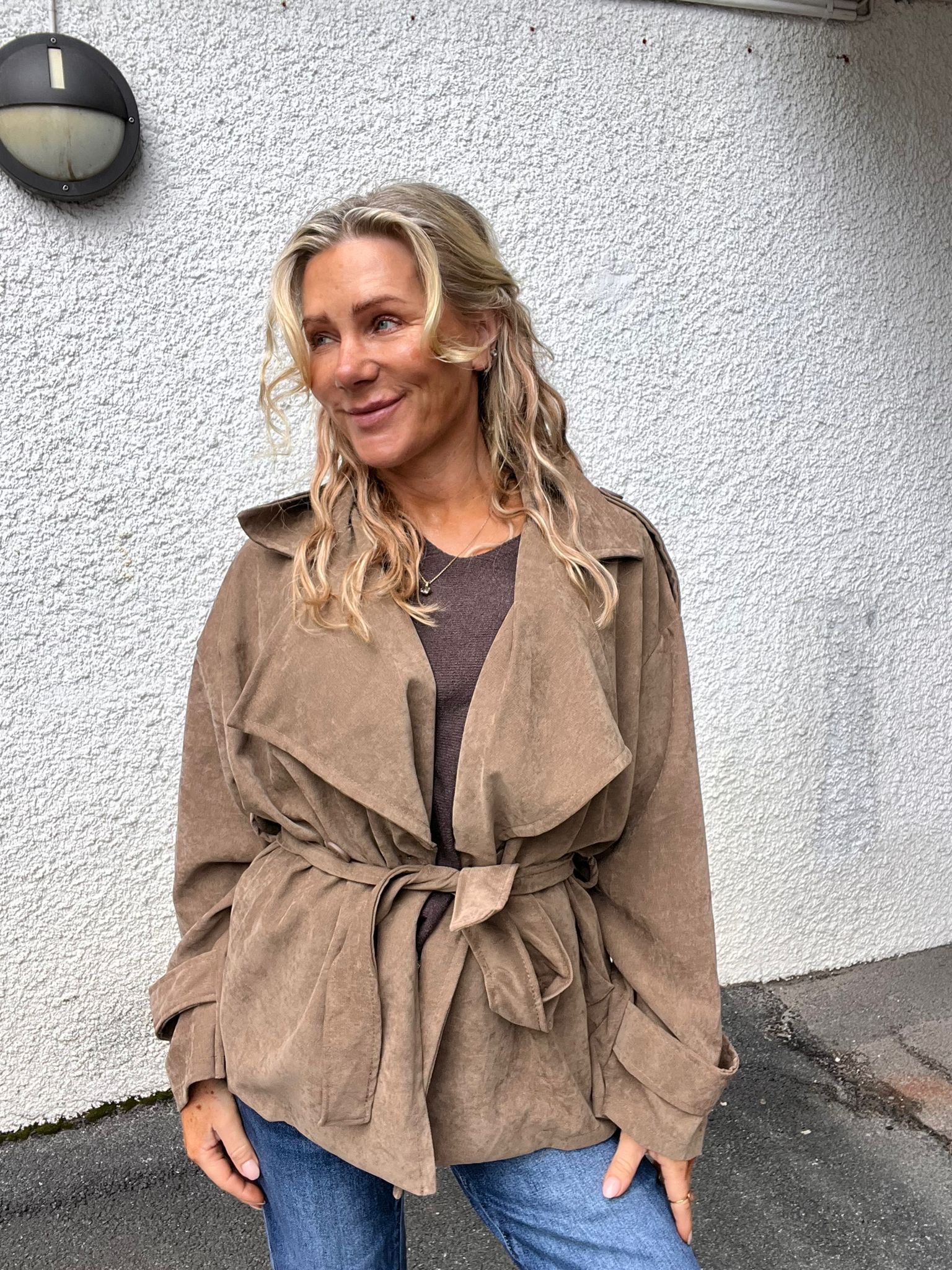 Kvinna med långt blont, vågigt hår står och ler medan hon tittar åt sidan, klädd i beige trenchcoat och jeans. Hon poserar mot en vit, grovputsad vägg utomhus med en rund vägglampa ovanför.