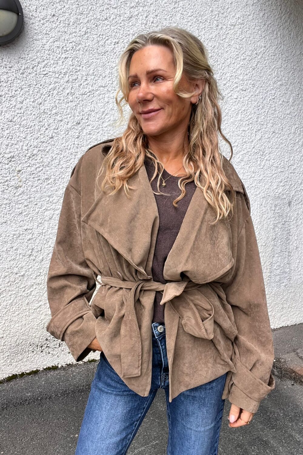 Blond kvinna står lutad mot en vit, grovputsad vägg och ler medan hon tittar åt sidan. Hon bär beige mockaliknande trenchjacka med knytbälte, brun topp, blå jeans och smycken; gatupromenadmiljö utomhus.
