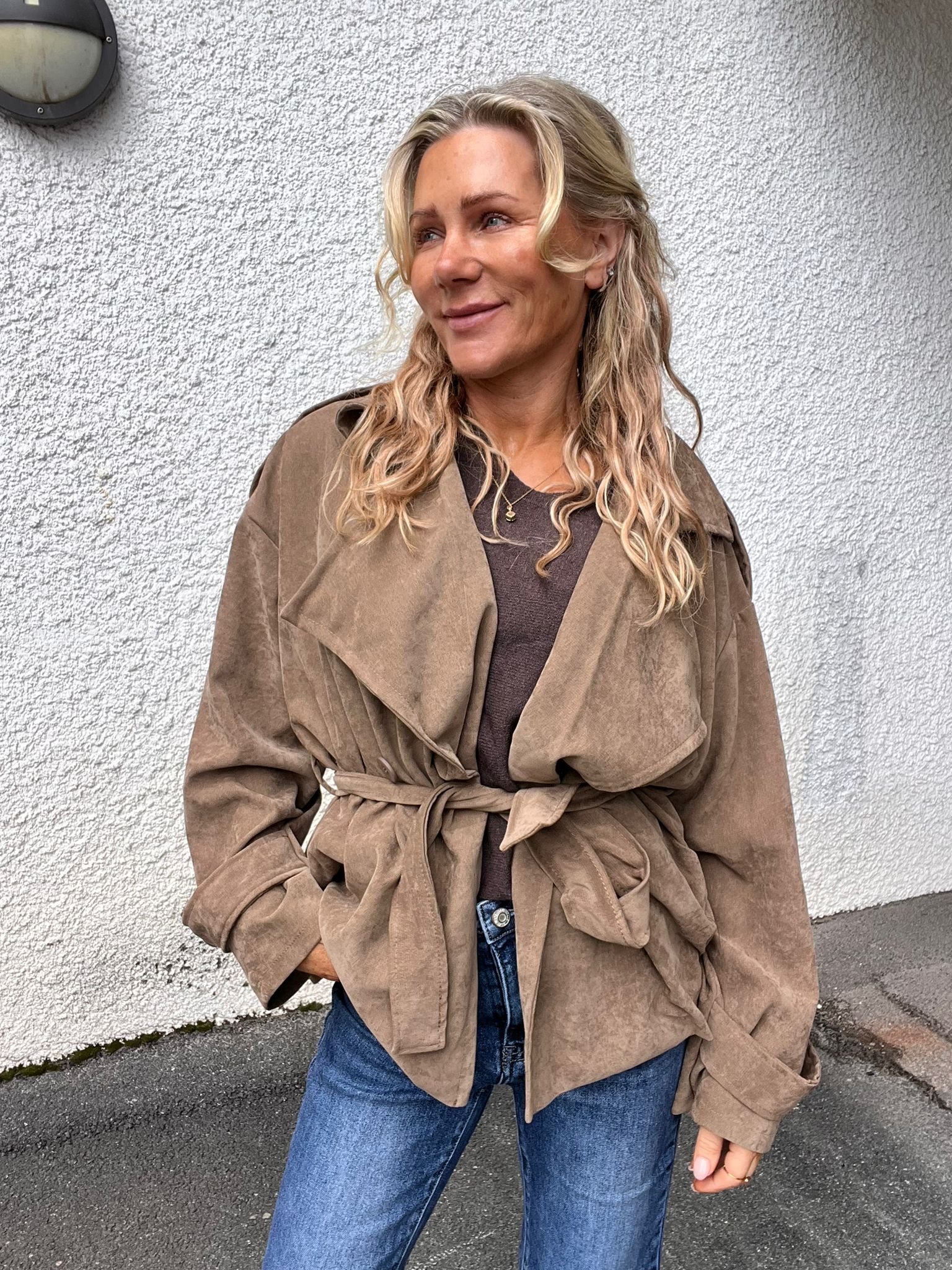 Blond kvinna står lutad mot en vit, grovputsad vägg och ler medan hon tittar åt sidan. Hon bär beige mockaliknande trenchjacka med knytbälte, brun topp, blå jeans och smycken; gatupromenadmiljö utomhus.