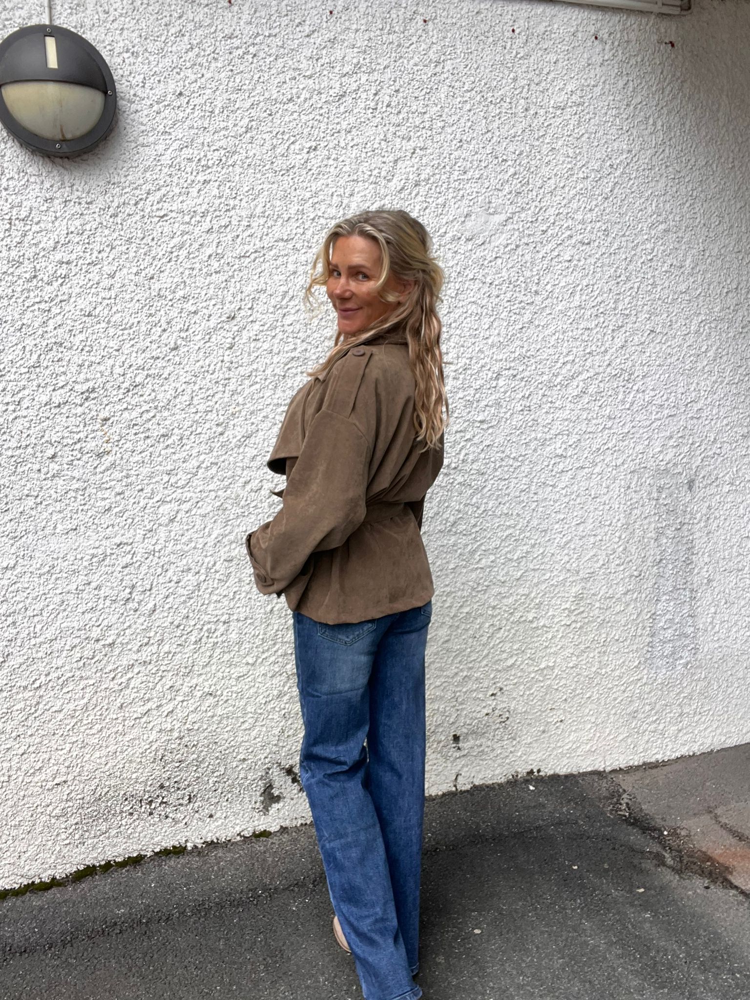 Kvinna med långt blont hår står i profil och vänder sig leende mot kameran. Hon bär brun jacka och blå jeans. Bakom henne syns en vit, grov putsad vägg med en rund vägglampa.