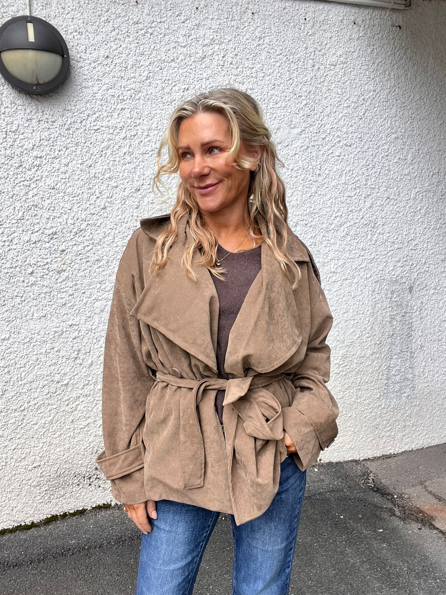 Kvinna med långt blont, vågigt hår står lutad mot en vit putsad vägg och ler medan hon tittar åt sidan. Hon bär beige trenchcoat med knytskärp och blå jeans; en rund vägglampa syns ovanför.