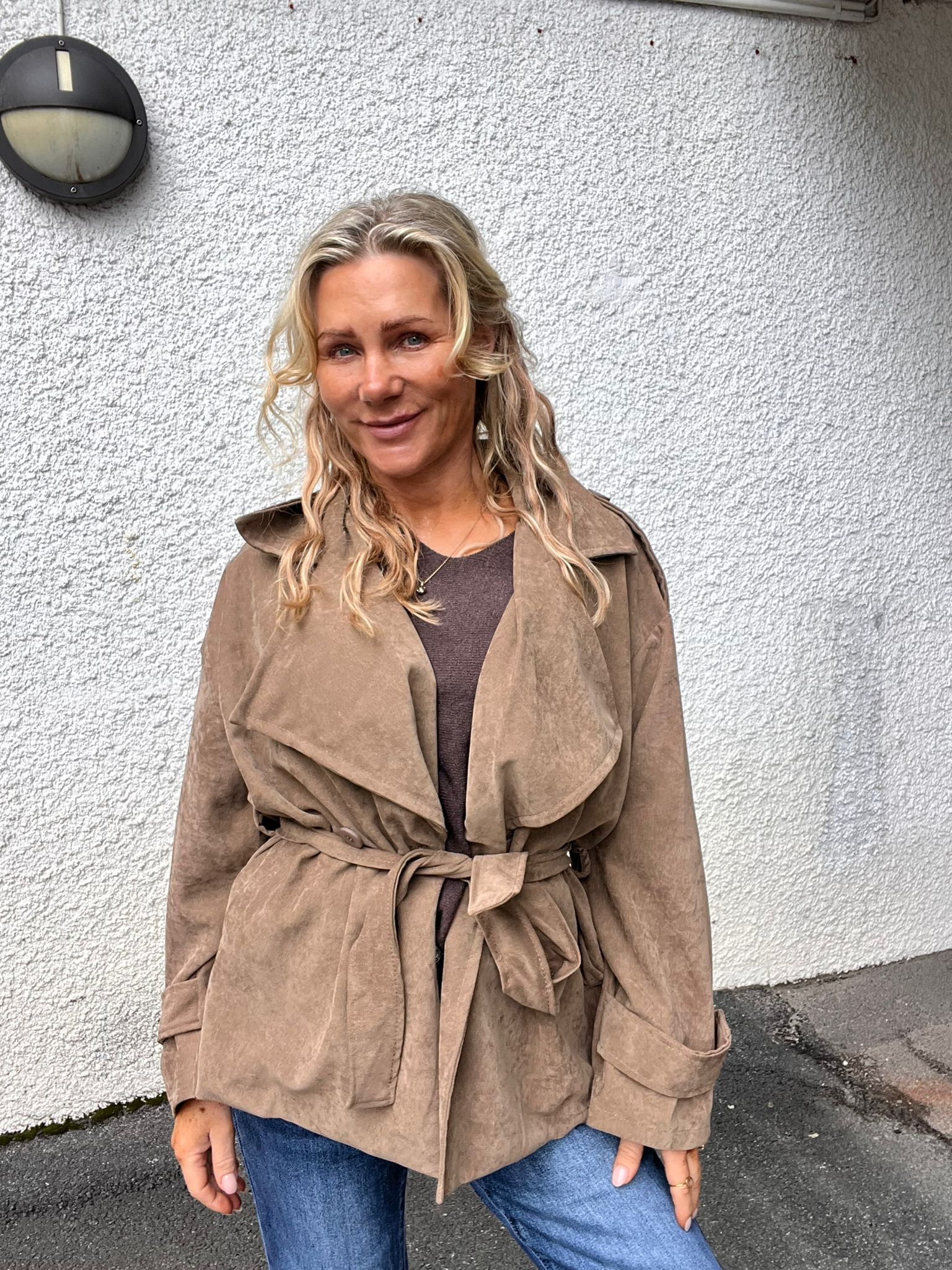 Kvinna med långt, vågigt blont hår står och ler mot kameran, klädd i brun mockajacka med skärp och blå jeans. Hon står utomhus framför en vit, grovputsad vägg med en rund vägglampa.