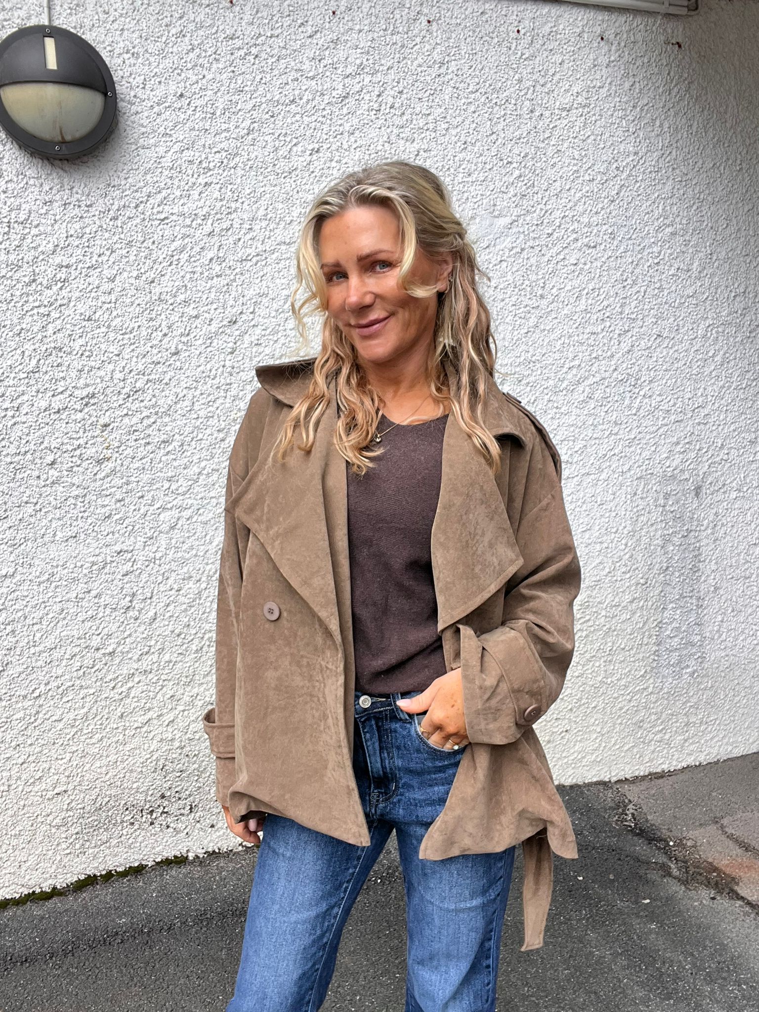Leende kvinna med långt blont hår står utomhus mot en vit, grovputsad vägg och tittar mot kameran. Hon bär beige trenchcoat, brun topp och blå jeans, med ena handen i fickan.