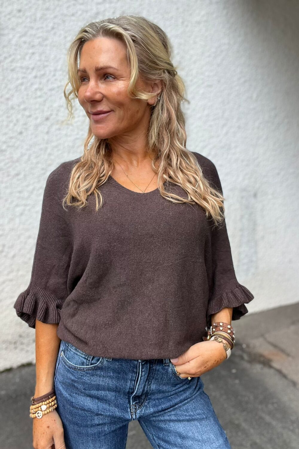 Blond kvinna står utomhus mot en ljus vägg och tittar åt vänster med ett litet leende. Hon bär brun stickad tröja med volangärmar och blå jeans, med armband och ringar på händerna.