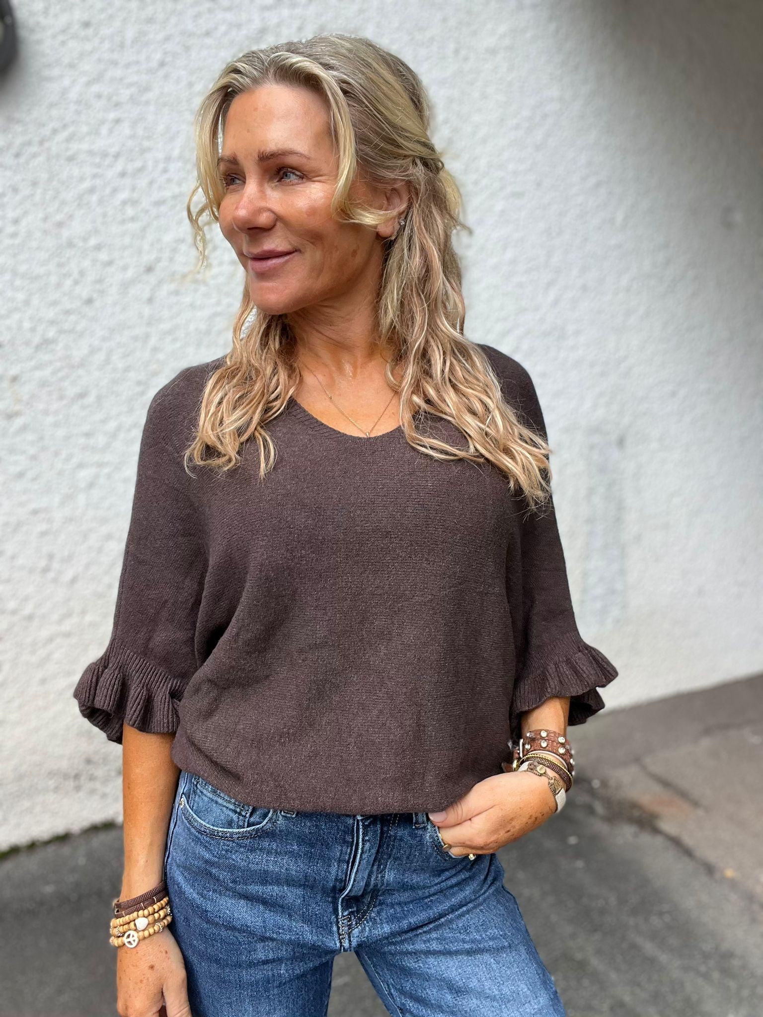 Blond kvinna står utomhus mot en ljus vägg och tittar åt vänster med ett litet leende. Hon bär brun stickad tröja med volangärmar och blå jeans, med armband och ringar på händerna.