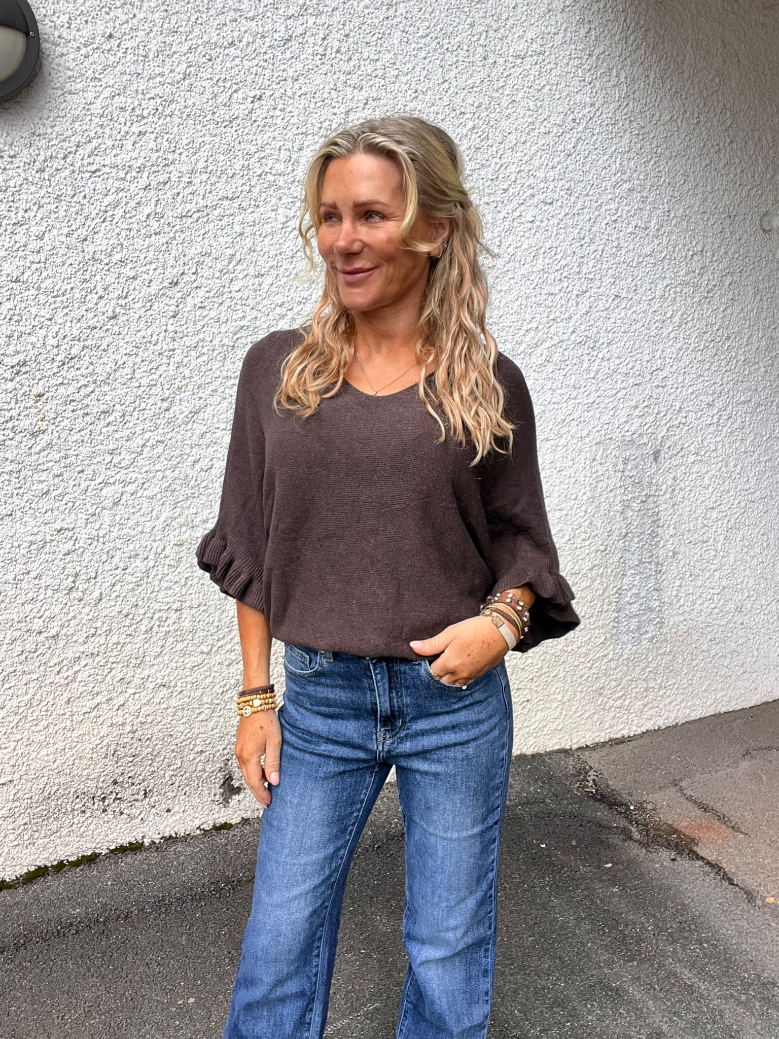 Blond kvinna står avslappnat och ler medan hon tittar åt sidan, med ena handen i jeansfickan. Hon bär mörk stickad tröja med volangärmar och flera armband, framför en vitputsad yttervägg utomhus.