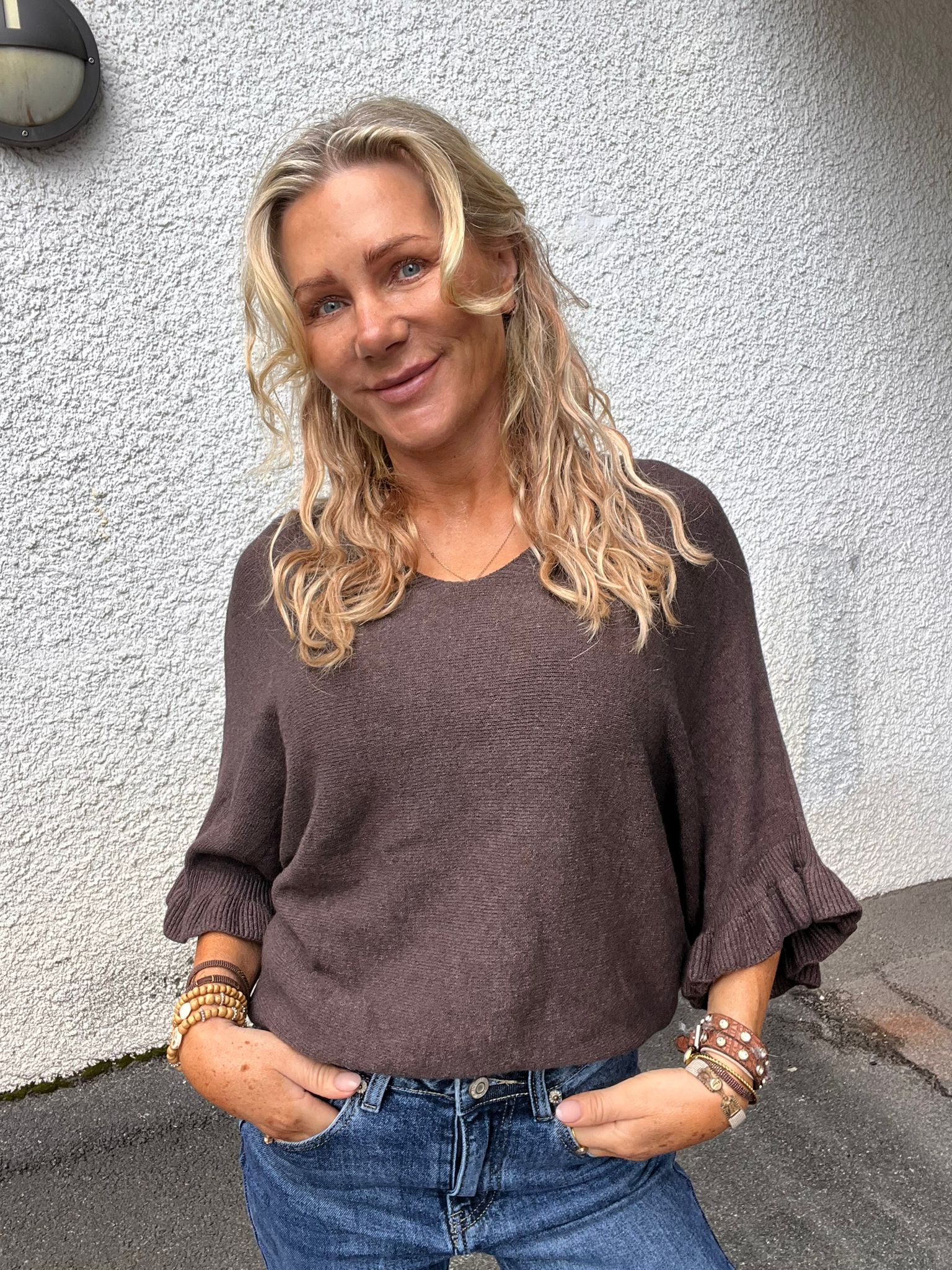 Leende kvinna med långt, vågigt blont hår står lutad mot en ljus, grovputsad vägg och tittar mot kameran. Hon har brun stickad tröja och jeans, händerna i fickorna och flera armband på handlederna.
