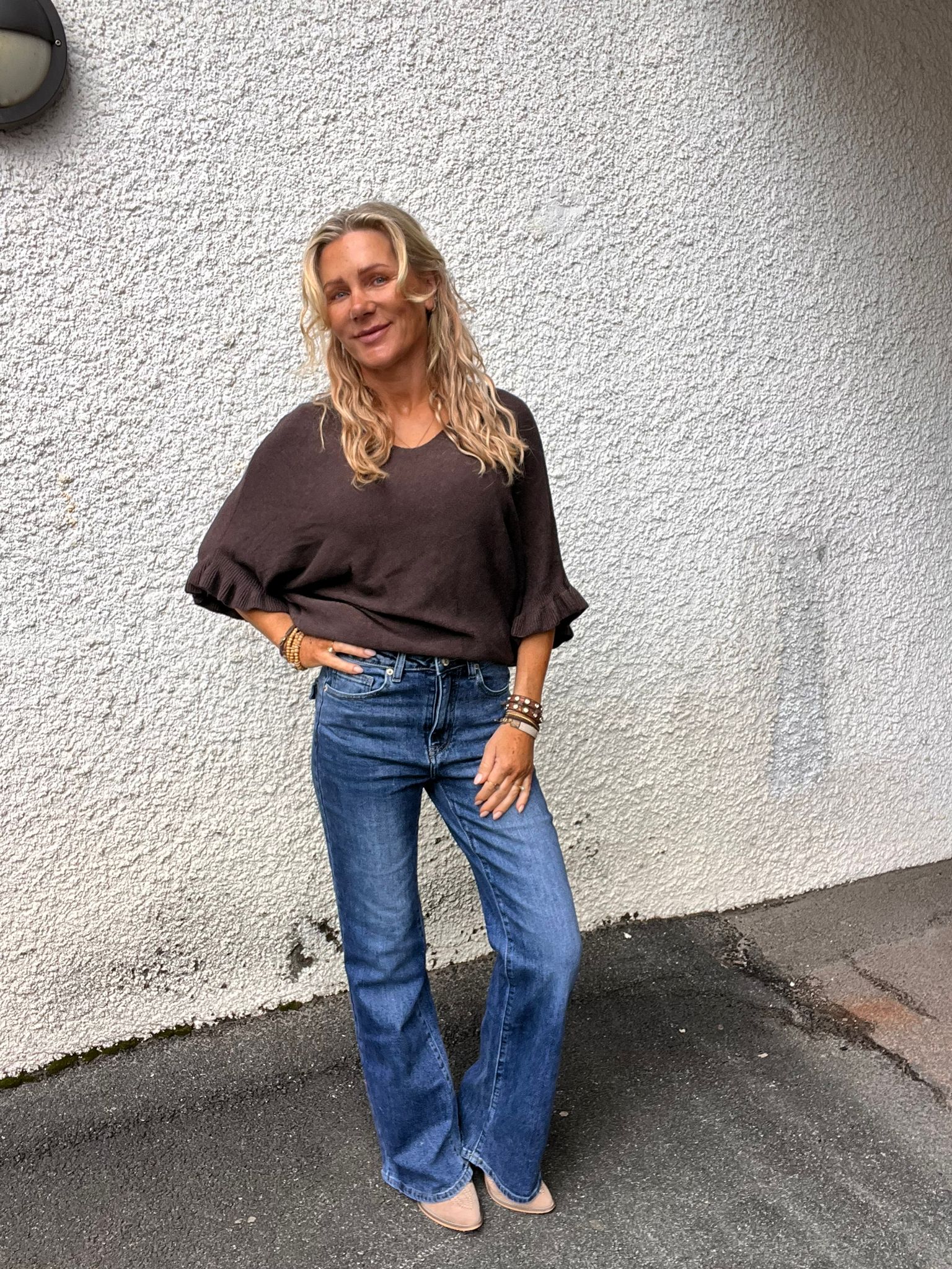 Kvinna med långt blont hår poserar avslappnat med en hand på höften och ler mot kameran. Hon bär mörk, löst sittande topp med volangärm, blå jeans och armband, stående utomhus mot grov vit vägg.