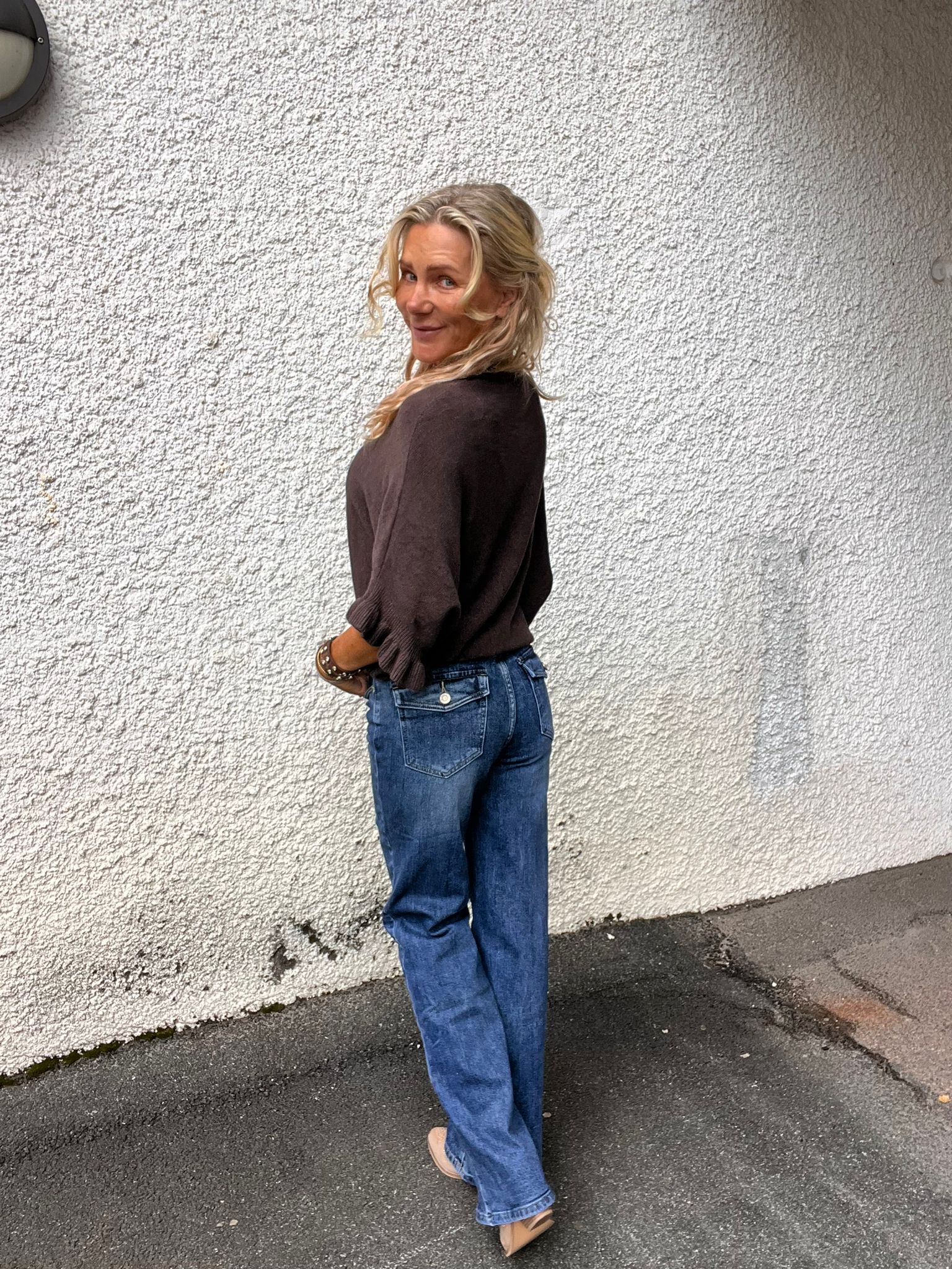Blond kvinna står i profil med händerna i fickorna och vänder sig om med ett leende mot kameran. Hon bär mörk långärmad tröja, blå jeans och beige klackskor, framför en vit, grovputsad vägg utomhus.