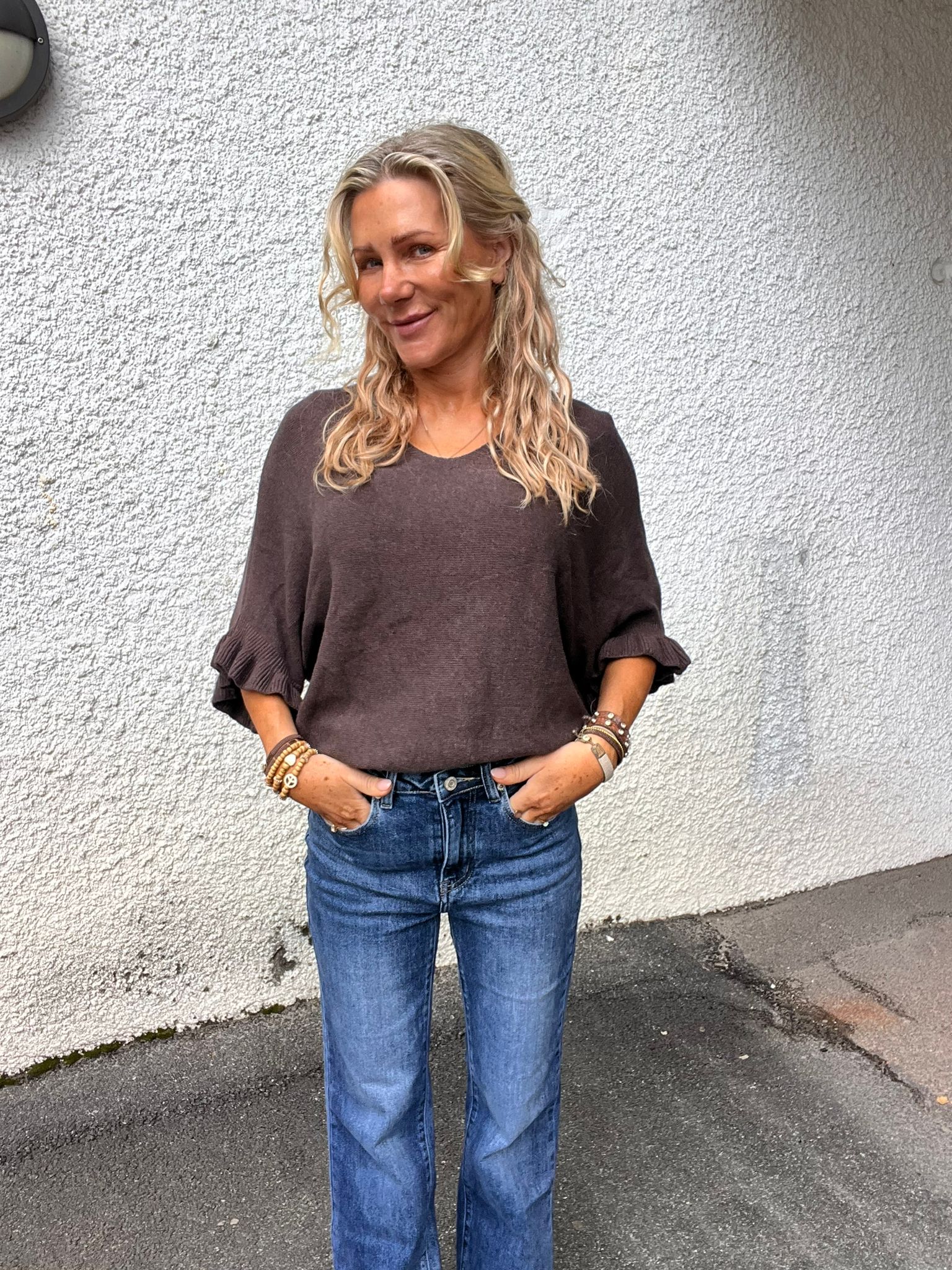 Blond kvinna står utomhus med händerna i jeansfickorna och ler lätt mot kameran. Hon bär brun topp med volangärmar och flera armband, framför en vit, grovputsad vägg på asfalt.