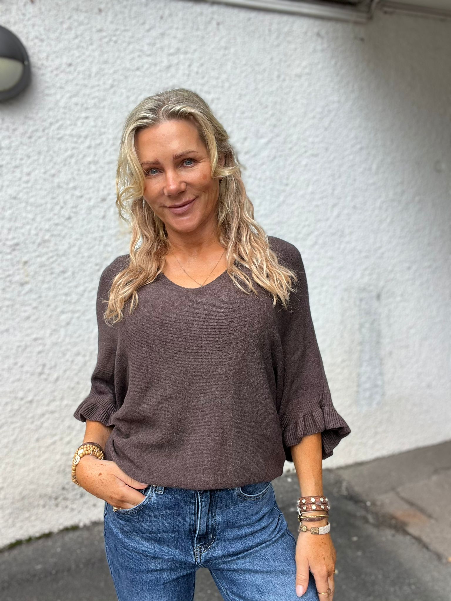 Blond kvinna står och ler mot kameran med ena handen i jeansfickan, iklädd brun stickad tröja med volangärmar och flera armband. Fotograferad utomhus framför en ljus, grovputsad vägg.