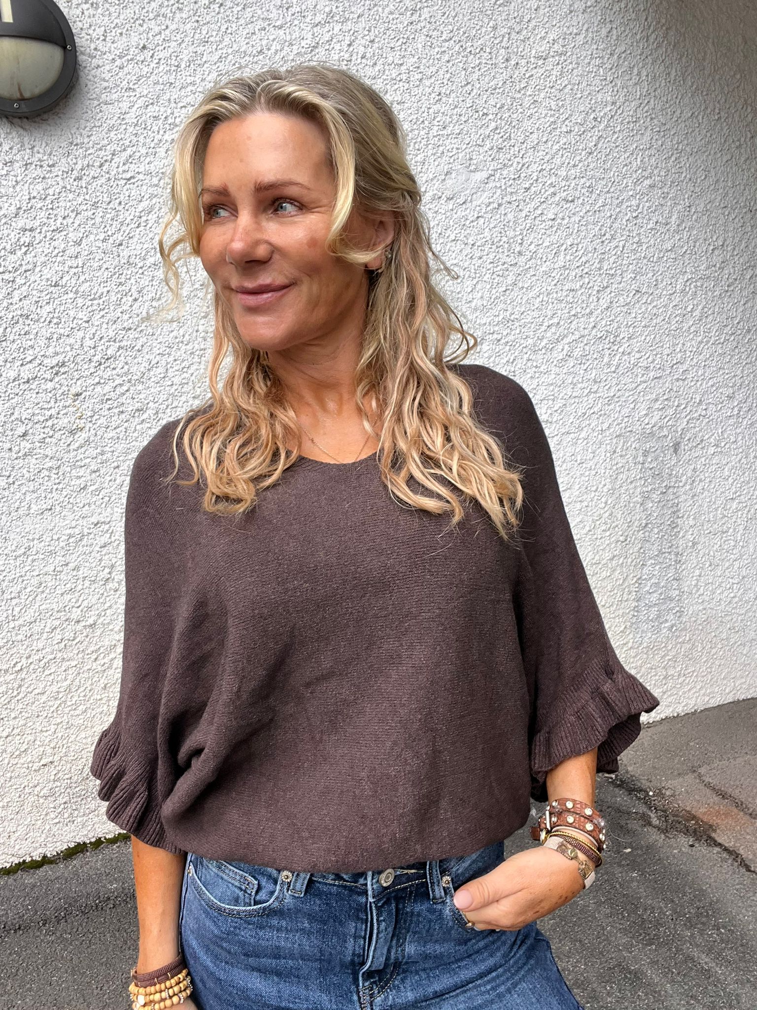Blond kvinna med lockigt hår står utomhus mot en vit putsad vägg och tittar åt vänster med ett svagt leende. Hon bär brun stickad tröja och blå jeans, med flera armband på handlederna.