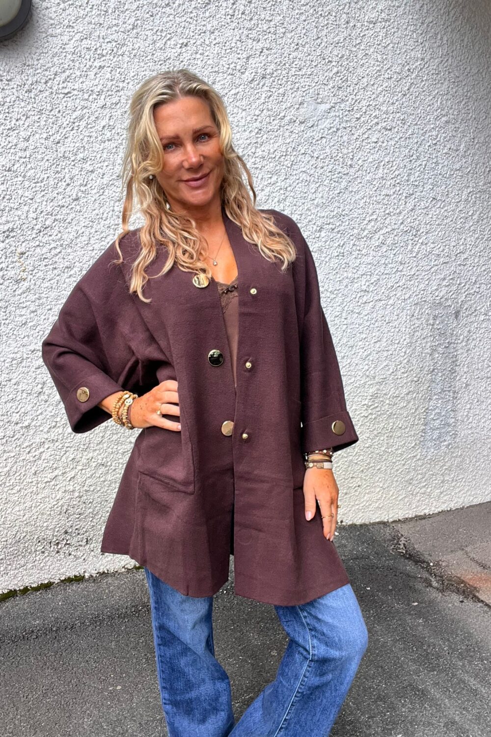 Blond kvinna poserar med handen på höften mot en vit, grovputsad vägg utomhus. Hon bär en mörklila lång jacka med stora guldfärgade knappar, blå jeans och flera armband.