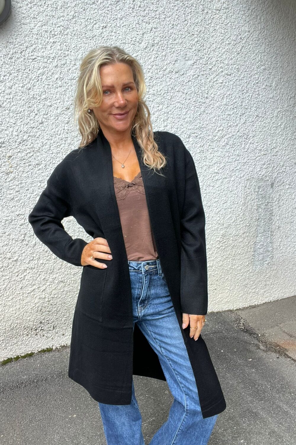 Blond kvinna står utomhus mot en vit, grovputsad vägg och ler mot kameran med handen på höften. Hon bär lång svart kofta, brun topp med spetskant och blå jeans.