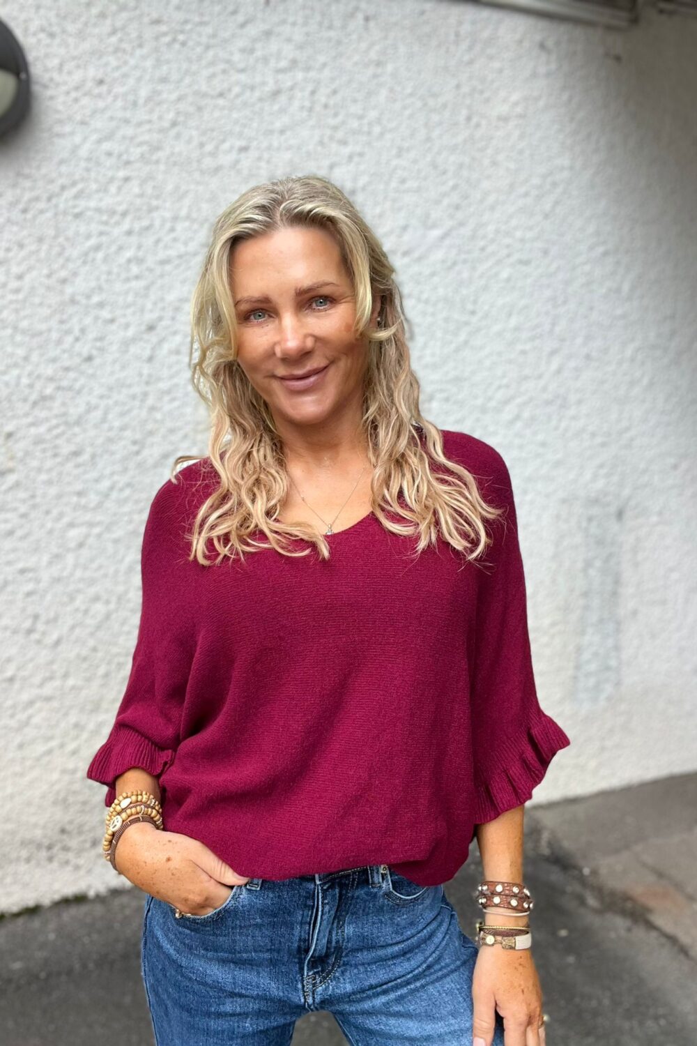 Leende kvinna med långt, vågigt blont hår står avslappnat med handen i jeansfickan. Hon bär vinröd stickad tröja med volangärmar och flera armband, framför en ljusgrå vägg utomhus.
