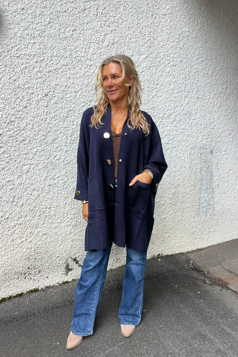 Kvinna med långt blont hår står avslappnat med ena handen i fickan och ler svagt. Hon bär mörkblå lång kofta med stora knappar, jeans och ljusa skor, mot en vit putsad vägg utomhus.