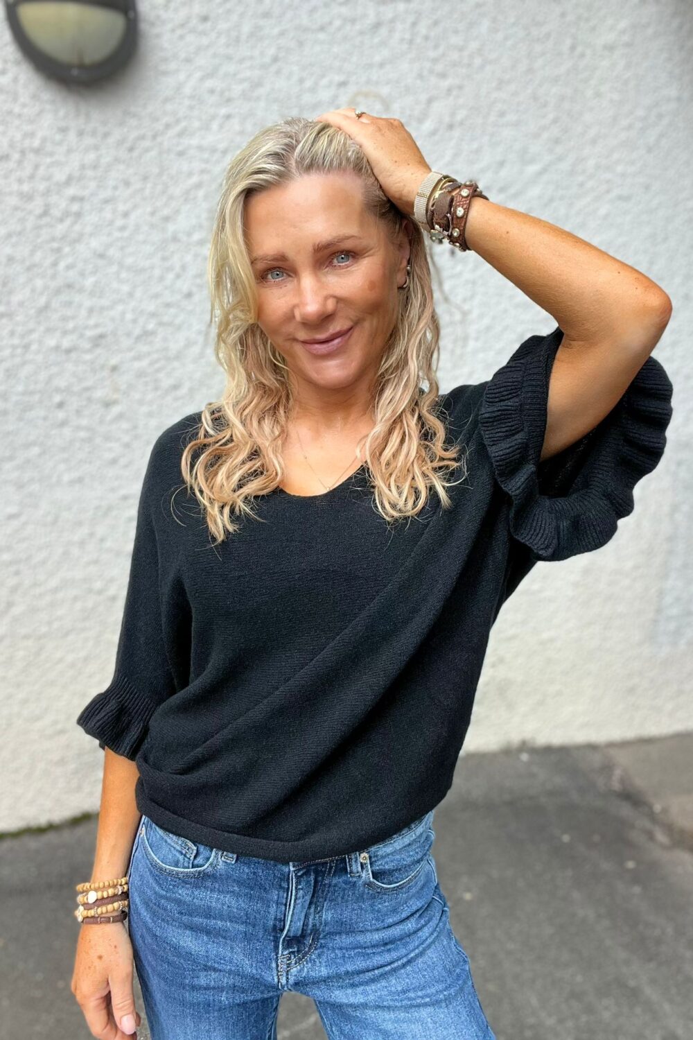 Blond kvinna står avslappnat och ler mot kameran med ena handen i håret. Hon bär svart stickad tröja med uppvikta ärmar, blå jeans och flera armband, framför en ljusgrå vägg utomhus.