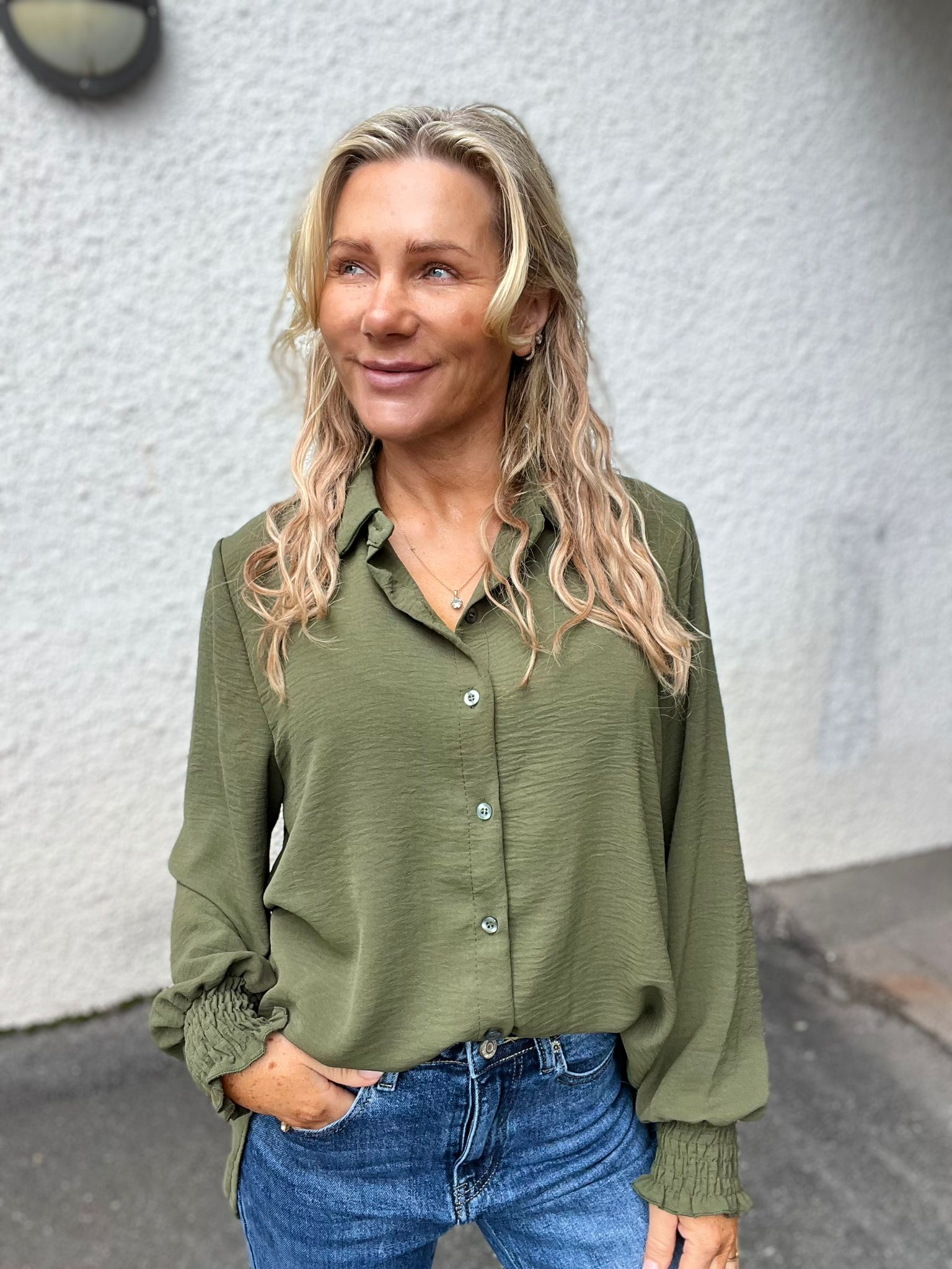 Leende kvinna med långt blont, vågigt hår står utomhus framför en ljusgrå vägg och tittar åt sidan. Hon har en olivgrön skjorta, blå jeans och ena handen i fickan.