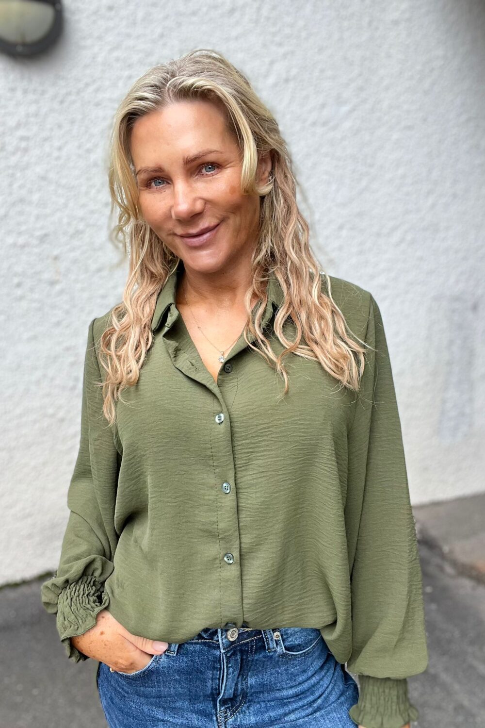 Blond kvinna med vågigt hår ler mot kameran och står avslappnat med ena handen i jeansfickan. Hon bär en olivgrön skjorta med rynkade manschetter och halsband, framför en ljus, slät vägg utomhus.