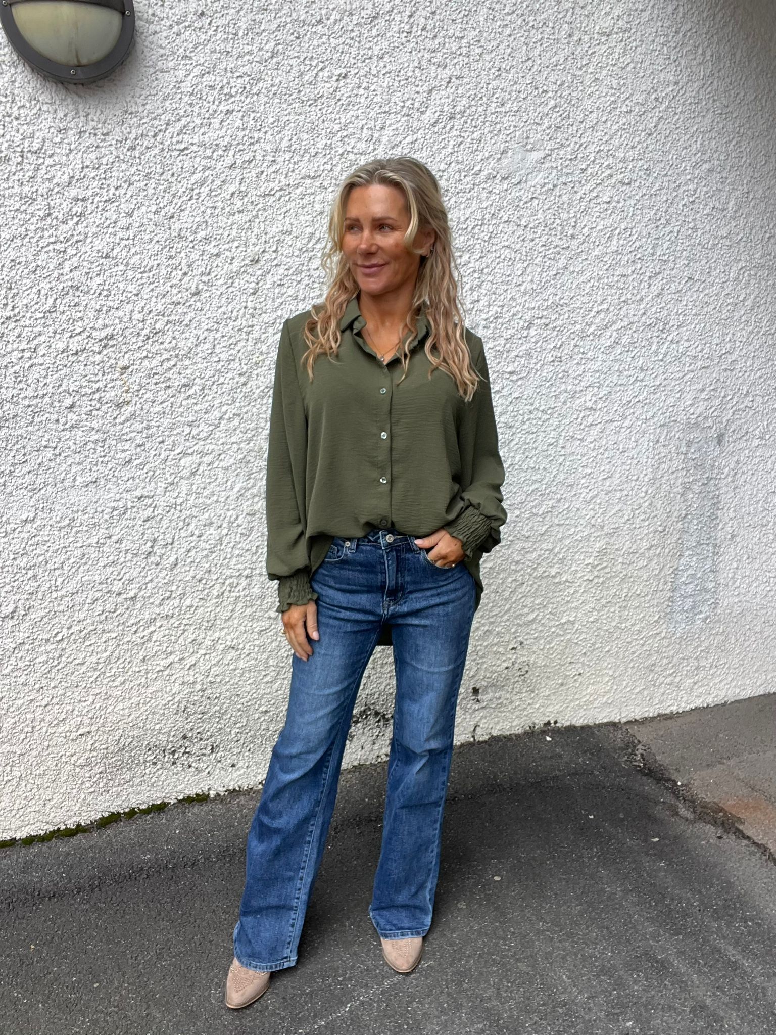 Kvinna med långt blont hår står avslappnat med ena handen i jeansfickan och tittar åt sidan. Hon bär olivgrön blus, blå jeans och beige skor, framför en vit putsad vägg med rund vägglampa.