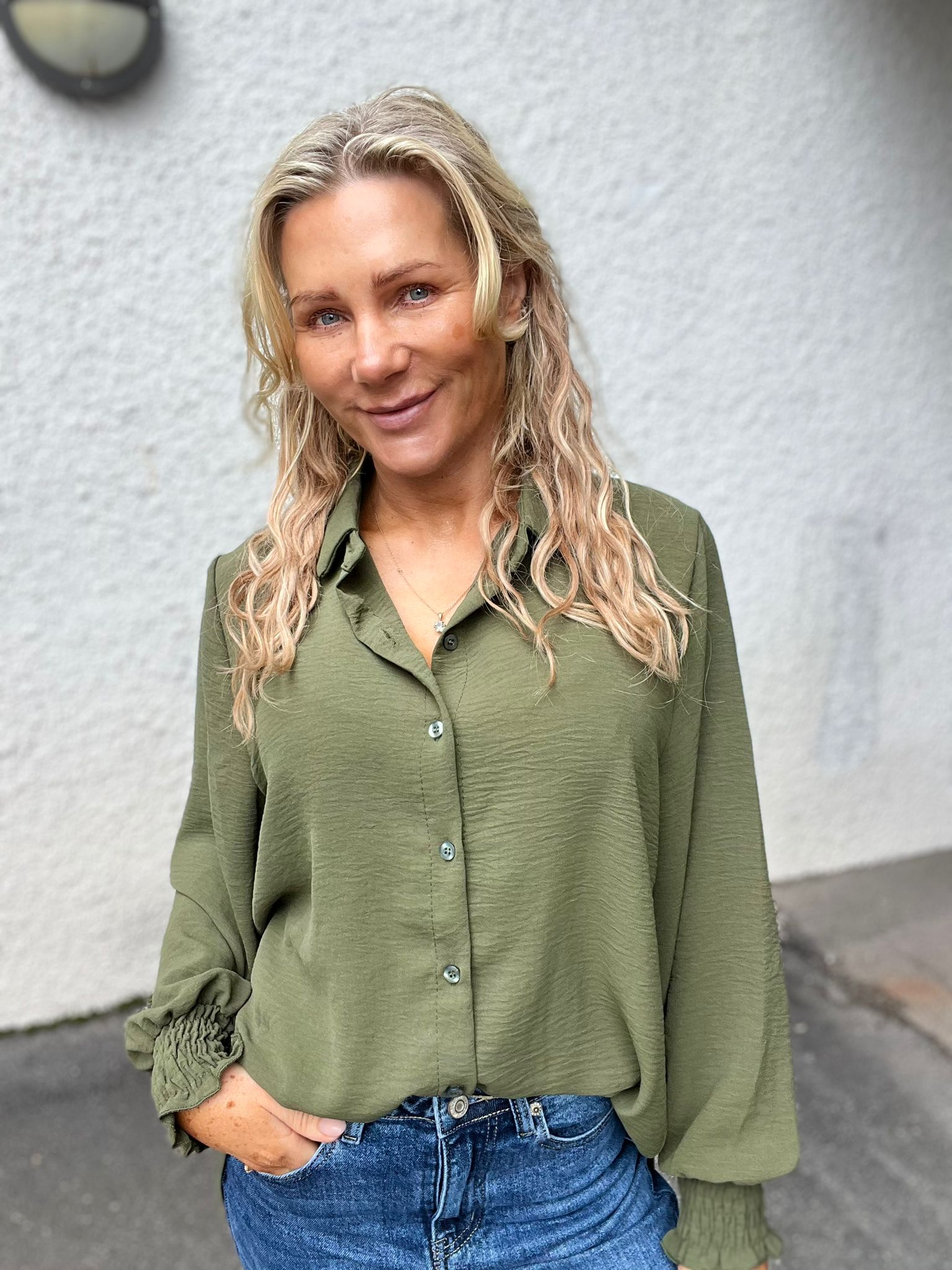 Blond kvinna med vågigt hår ler mot kameran och står avslappnat med ena handen i jeansfickan. Hon bär en olivgrön skjorta med rynkade manschetter och halsband, framför en ljus, slät vägg utomhus.