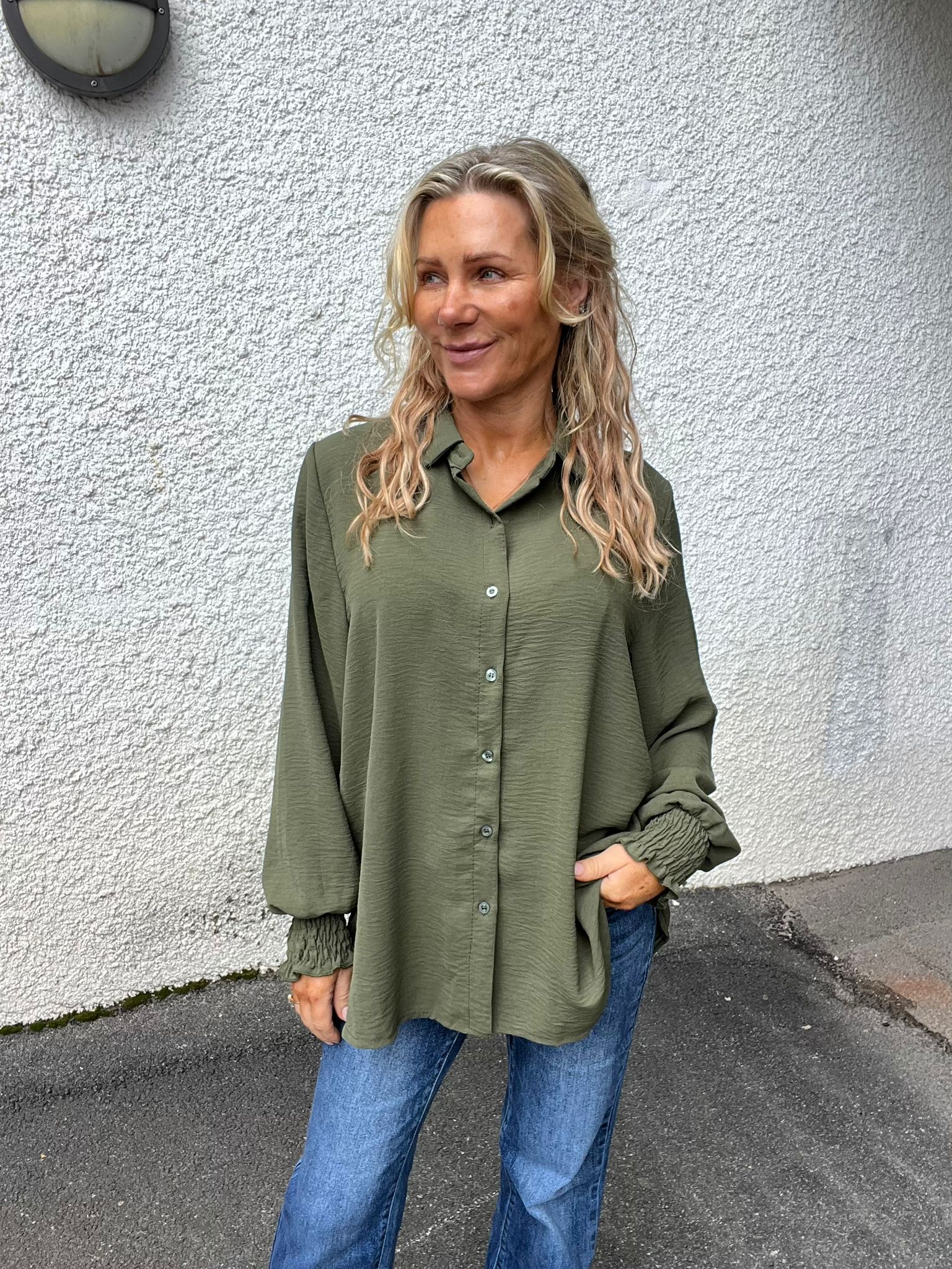 Kvinna med långt blont, vågigt hår står avslappnat och tittar åt sidan, med ena handen i jeansfickan. Hon bär en olivgrön, långärmad skjorta med knappar och står framför en vit putsad vägg utomhus.