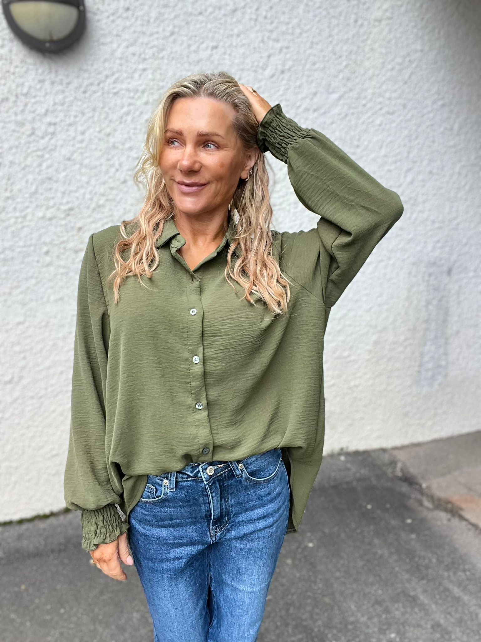 Kvinna med långt blont hår står utomhus framför en ljus, grovputsad vägg, ler och tittar åt sidan medan hon håller ena handen bakom huvudet. Hon bär grön blus och blå jeans.
