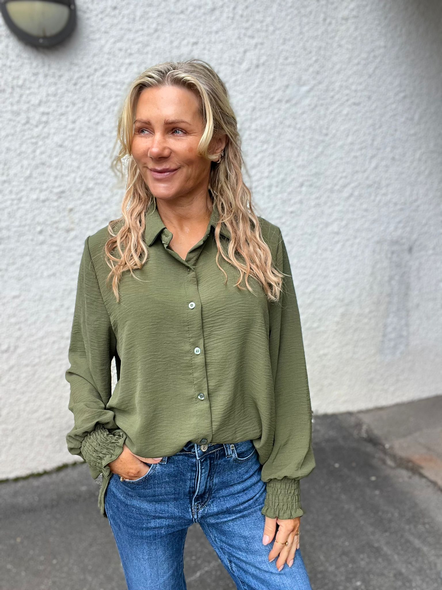 Blond kvinna står avslappnat utomhus med ena handen i jeansfickan och ler medan hon tittar åt sidan. Hon bär en olivgrön, knappad blus med rynkade muddar. Bakgrund: ljus putsad vägg och asfalt.