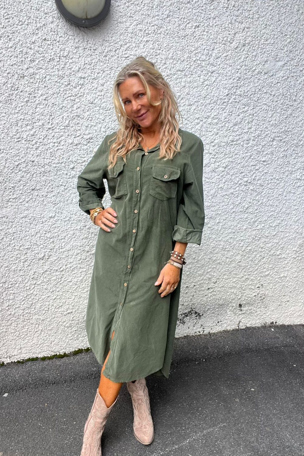 Blond kvinna står och poserar med ena handen på höften framför en vit, grovputsad vägg. Hon bär en olivgrön skjortklänning och ljusa cowboyboots samt flera armband; utomhus på asfalt.