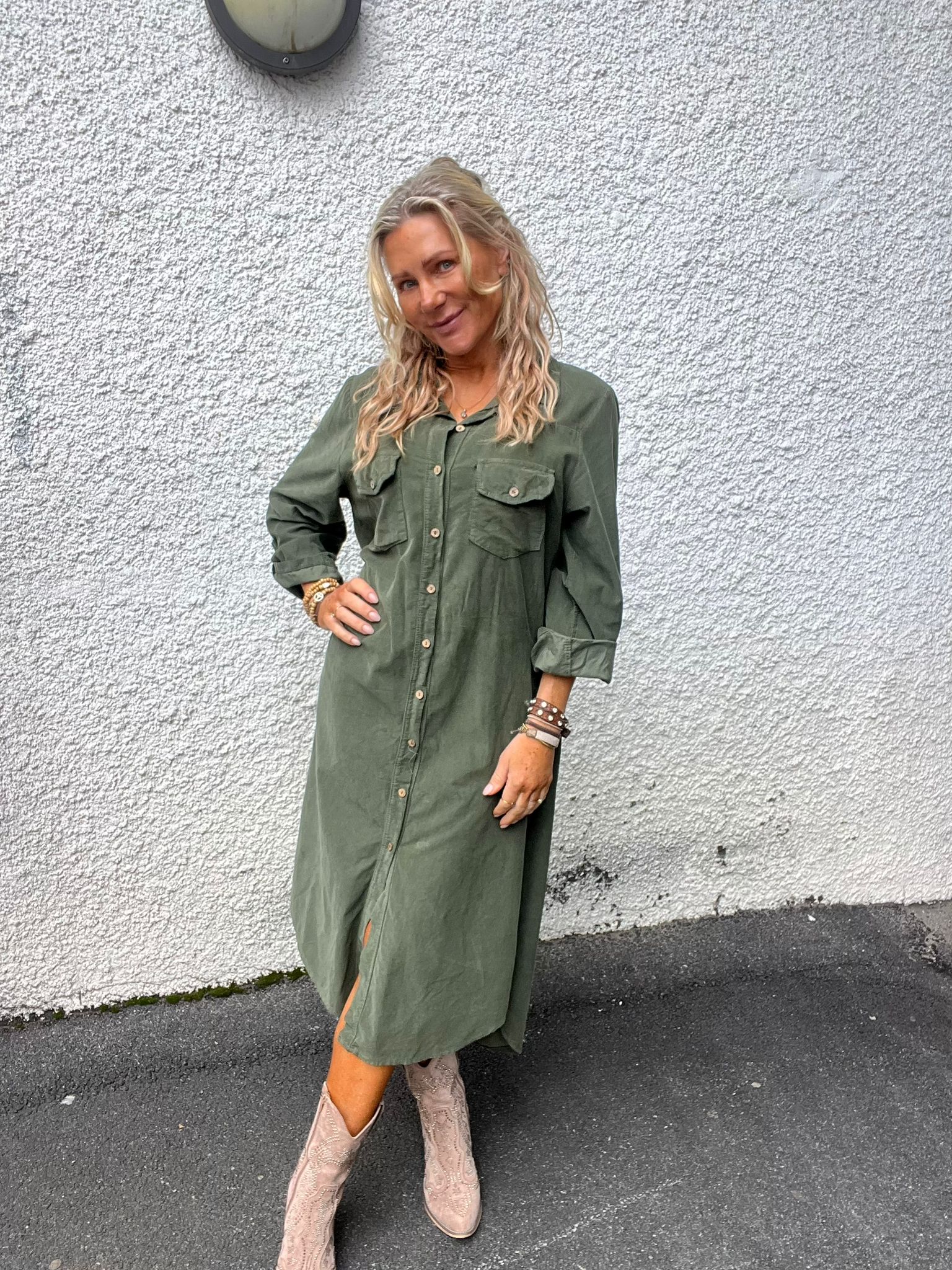 Blond kvinna står och poserar med ena handen på höften framför en vit, grovputsad vägg. Hon bär en olivgrön skjortklänning och ljusa cowboyboots samt flera armband; utomhus på asfalt.