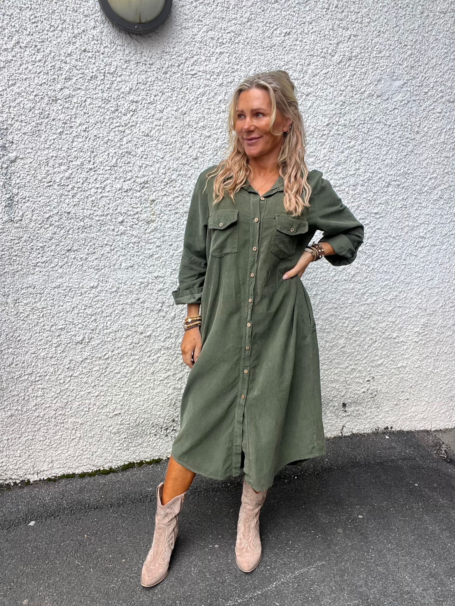 Kvinna i grön skjortklänning poserar med ena handen på höften och blicken åt sidan. Hon står mot en vit, grovputsad vägg med vägglampa ovanför och bär ljusa cowboyboots samt armband.