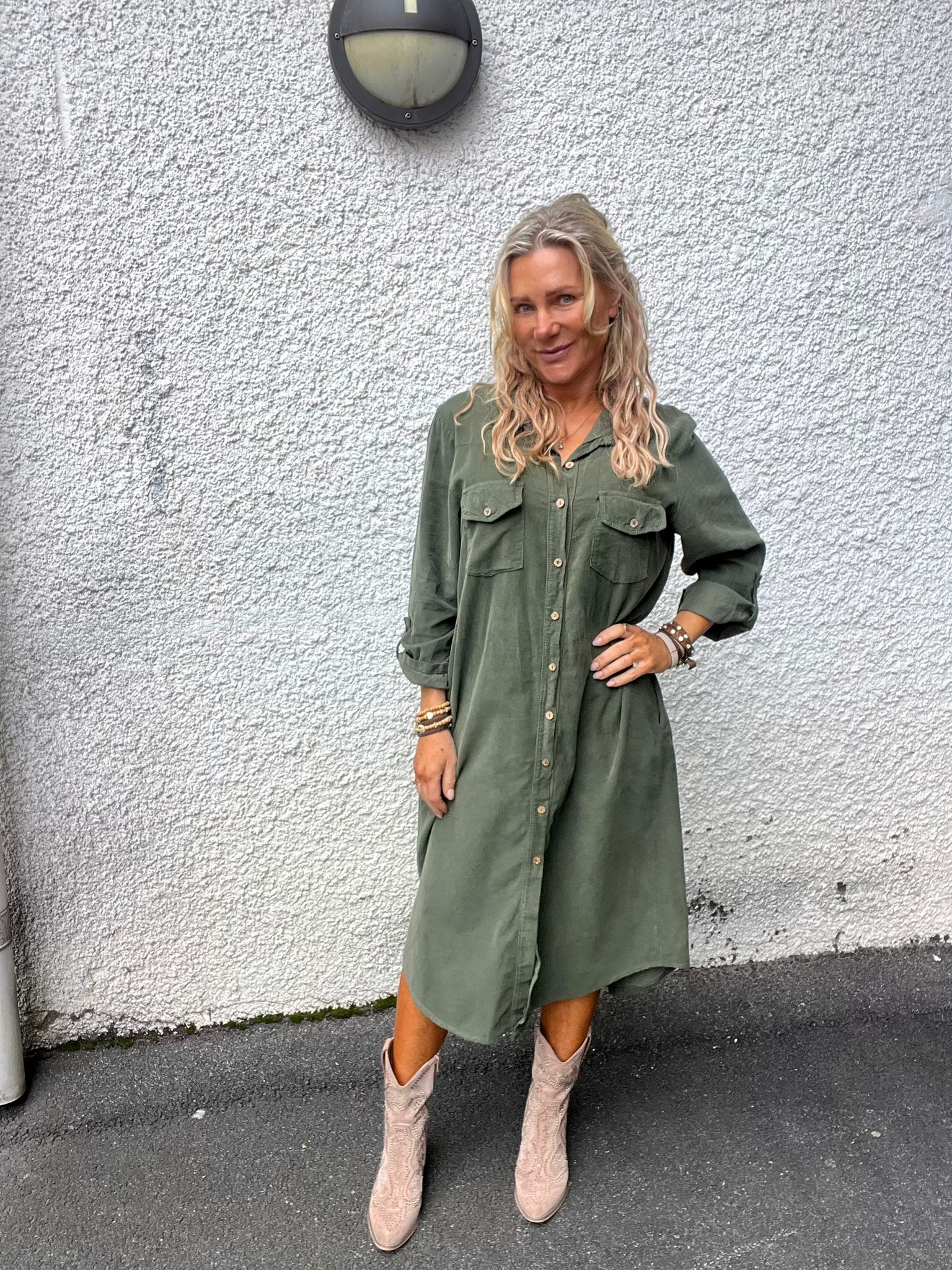 Kvinna med långt blont hår poserar med ena handen på höften framför en vit, grovputsad vägg. Hon bär grön skjortklänning med knappar, armband och ljusa cowboyboots; en rund vägglampa syns ovanför.