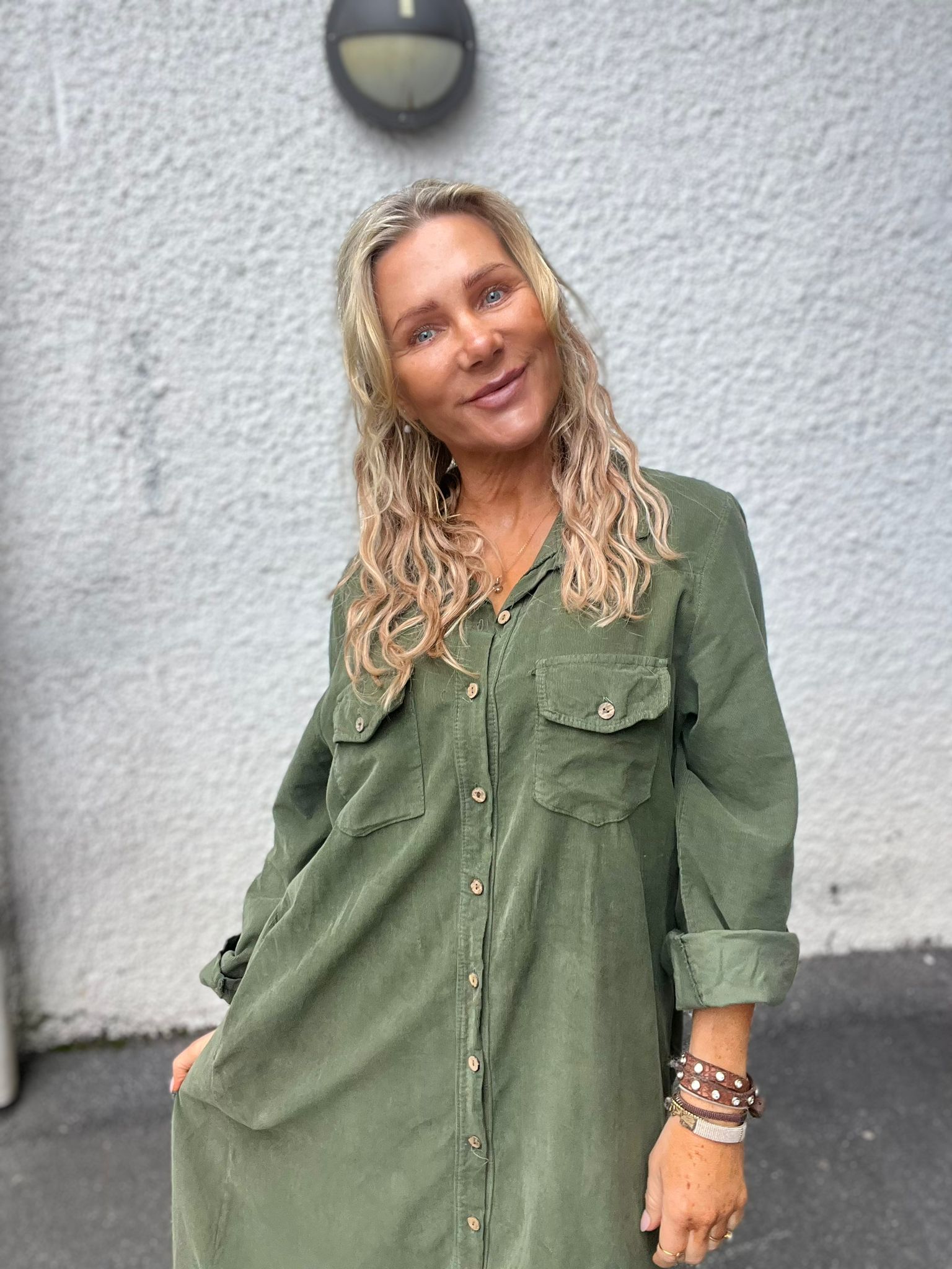 Blond kvinna i grön, lång skjortklänning lutar sig lätt och ler mot kameran. Hon står utomhus framför en ljusgrå putsad vägg med en rund vägglampa ovanför. Armband syns på handleden.