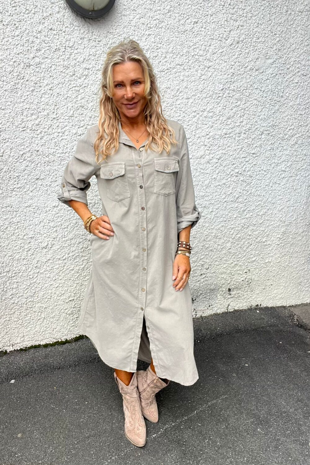 Leende kvinna står med ena handen på höften framför en vit, grovputsad vägg. Hon bär en ljusgrå skjortklänning med uppkavlade ärmar, flera armband och ljusa cowboyboots på mörk asfalt.