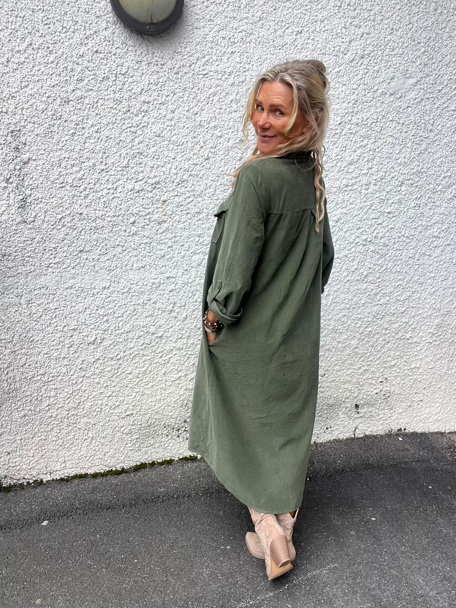 Blond kvinna i lång olivgrön skjortklänning står med händerna i fickorna och ser över axeln mot kameran, framför en vit grovputsad vägg; hon bär beige boots och armband på en asfalterad gång.