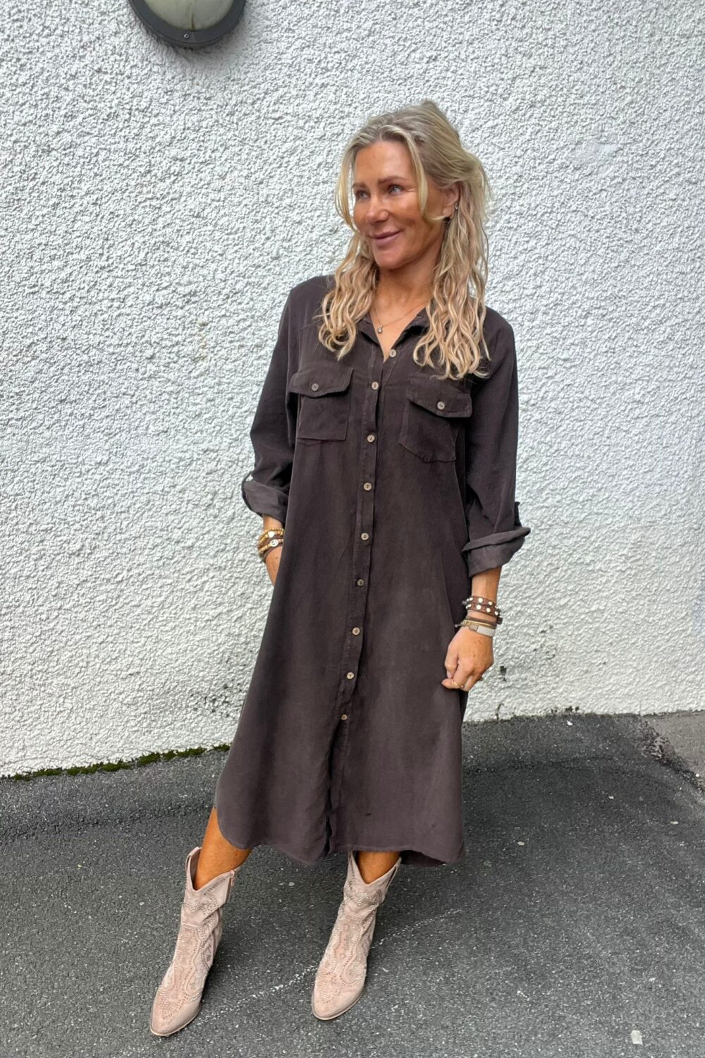 Kvinna med långt blont, lockigt hår står lutad mot en vit, grovputsad vägg och ler åt sidan. Hon bär en mörkgrå skjortklänning med knappar, armband och ljusa cowboyboots på asfalt utomhus.