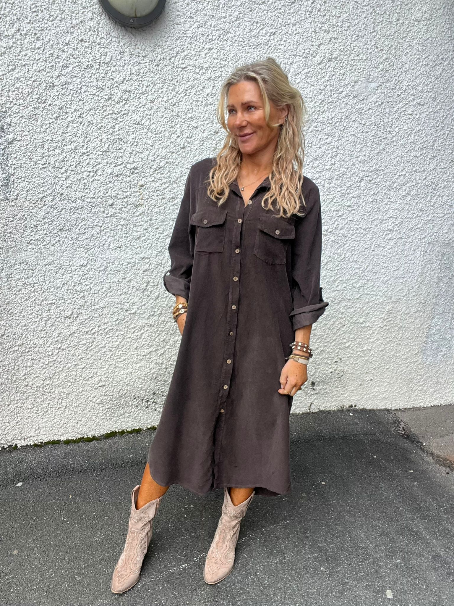 Kvinna med långt blont, lockigt hår står lutad mot en vit, grovputsad vägg och ler åt sidan. Hon bär en mörkgrå skjortklänning med knappar, armband och ljusa cowboyboots på asfalt utomhus.